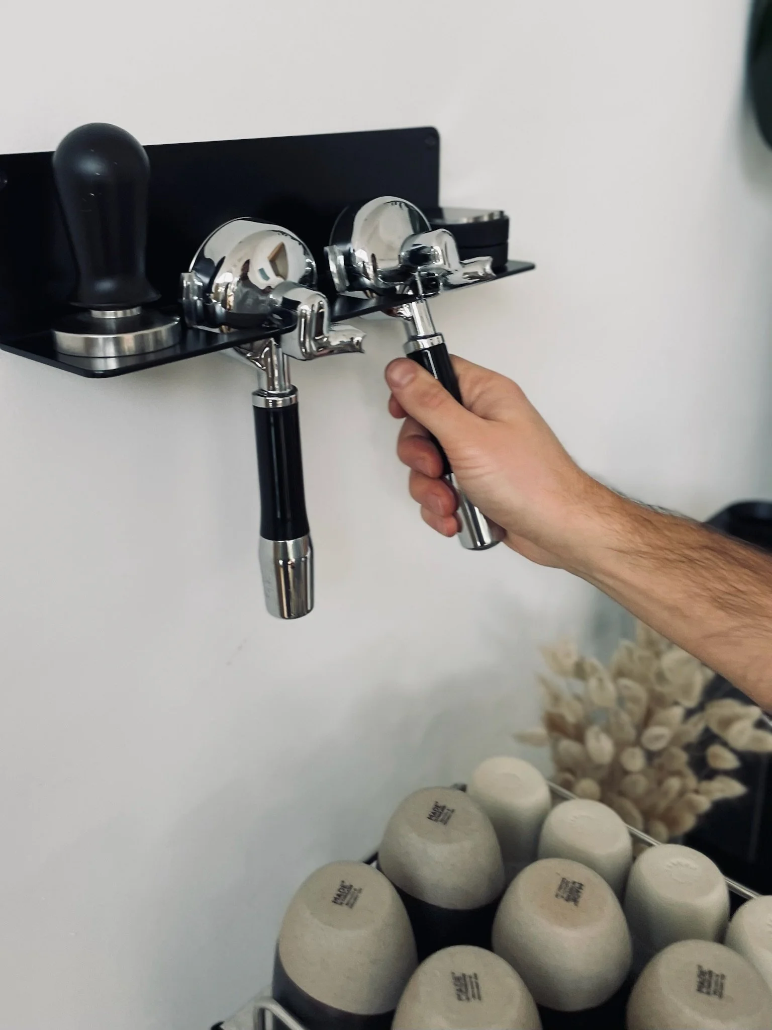 Hand, die einen Siebträger von einem Baristaregal nimmt, mit umgedrehten Bechern im Vordergrund und einem Tamper daneben.