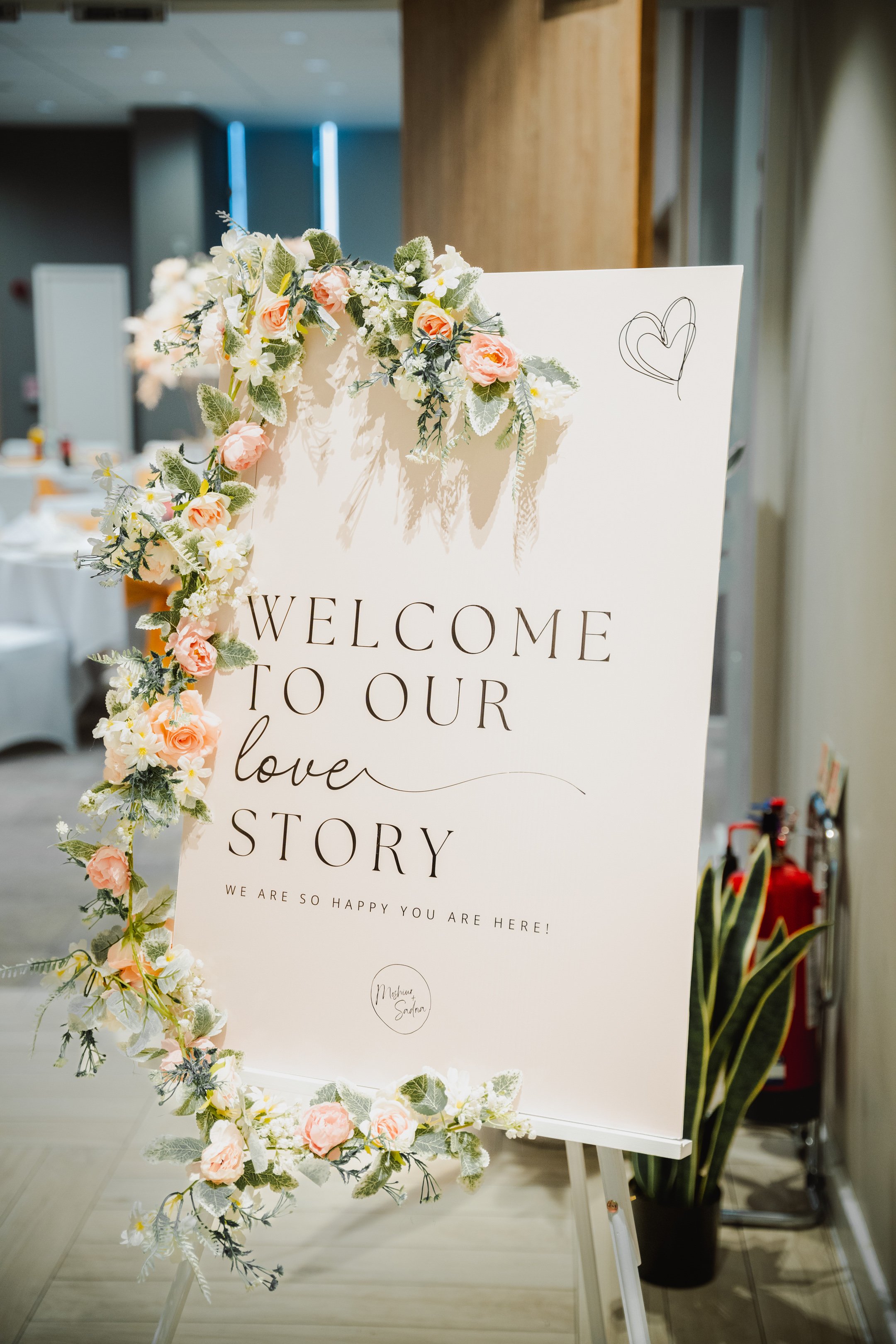 Wedding welcome sign decorated with pink and white flowers and green foliage, reading 'Welcome to our love story' with smaller text below, in an indoor venue.