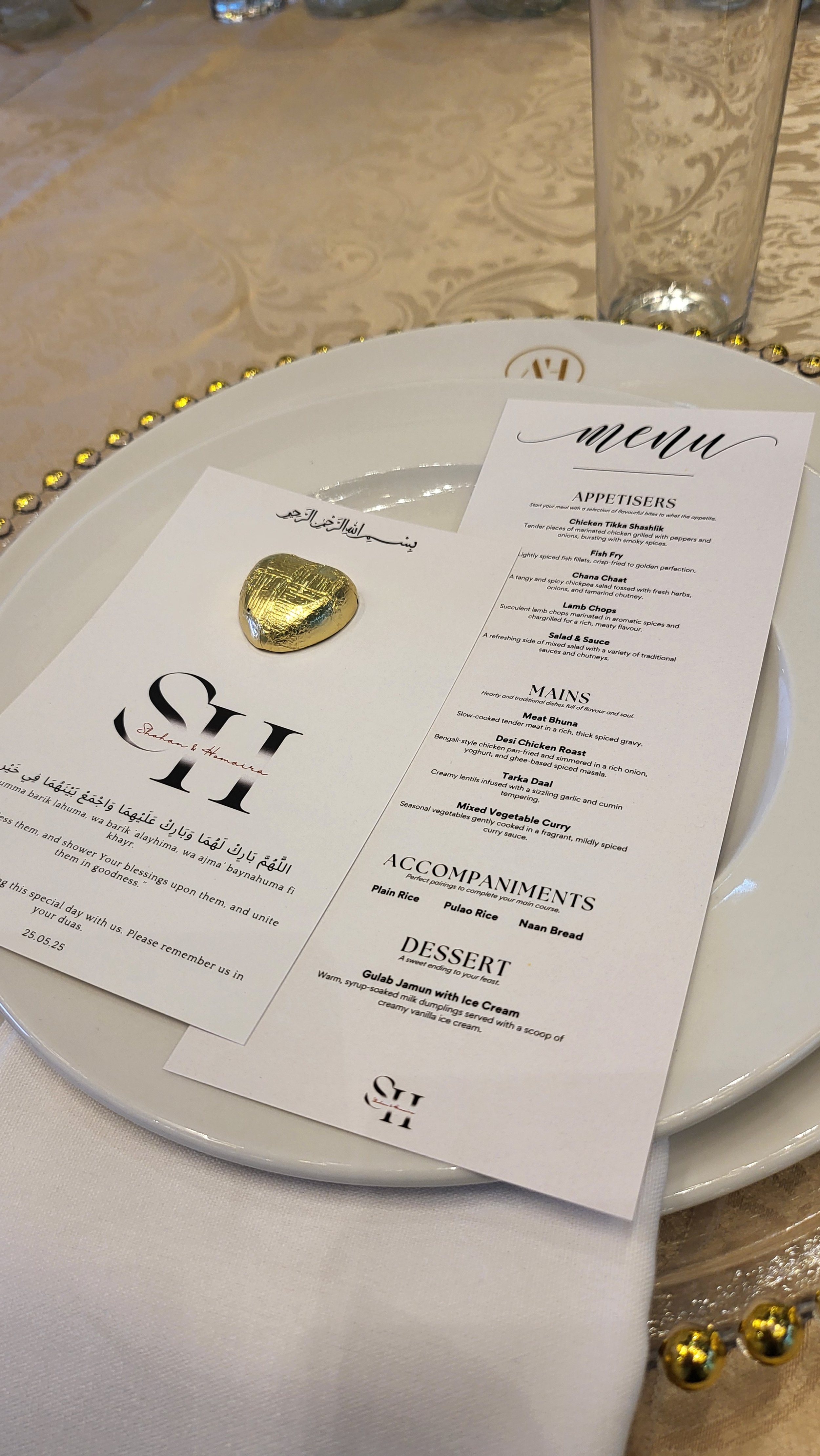 A fancy table setting with a menu, a white plate with a gold heart-shaped pin, and a drinking glass, on a beige tablecloth with gold embellishments.