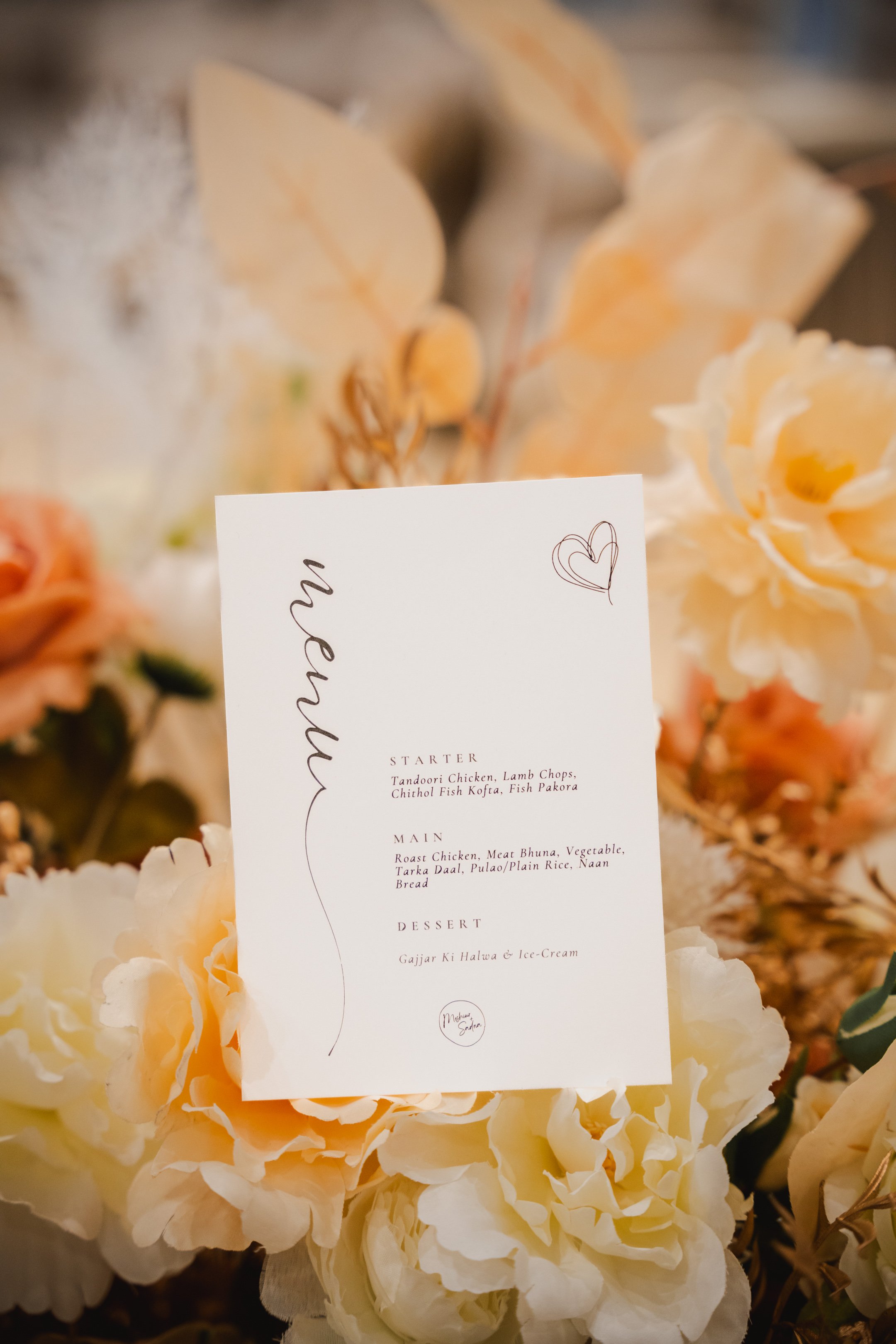 A wedding menu card on a bed of cream-colored flowers with a heart doodle in the top right corner.