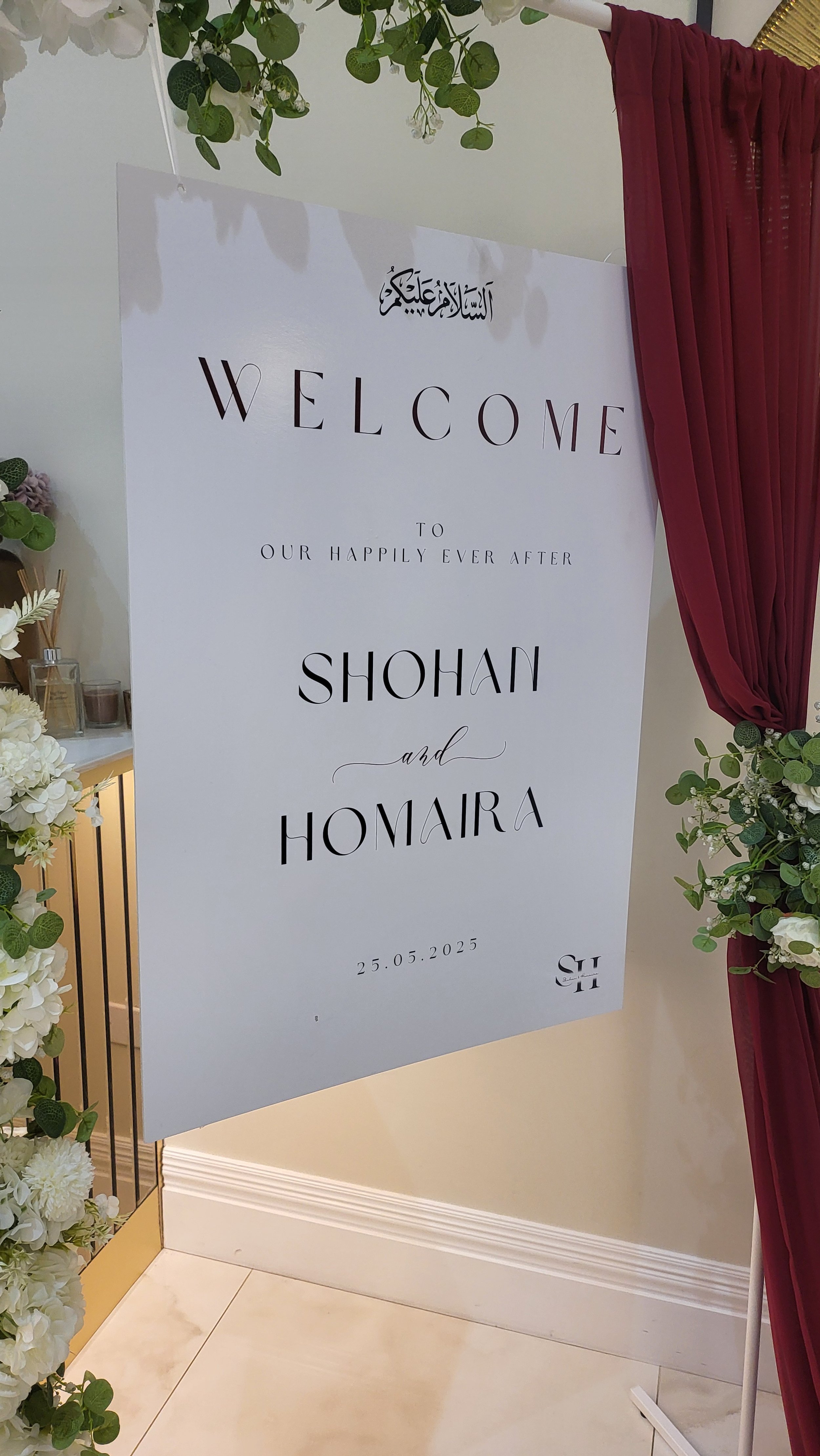 Wedding welcome sign with floral decorations and red curtains, displaying the date 25.05.2025.