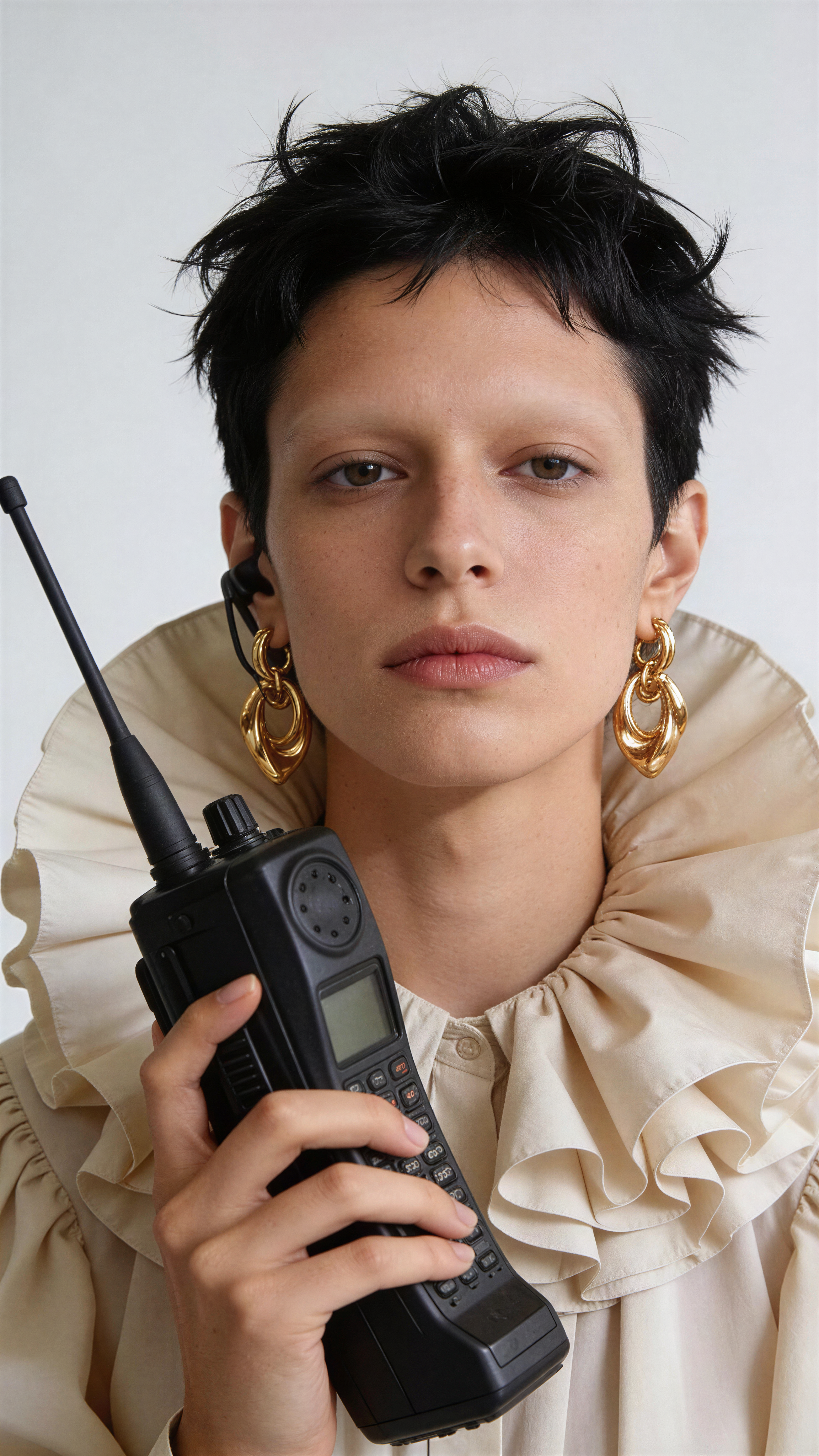 Porträt einer jungen Frau mit schwarzen, kurzen, zerzausten Haaren, goldenen Ohrringen, hellbeigem Rüschen-Hemd und einem Funkgerät in der Hand.