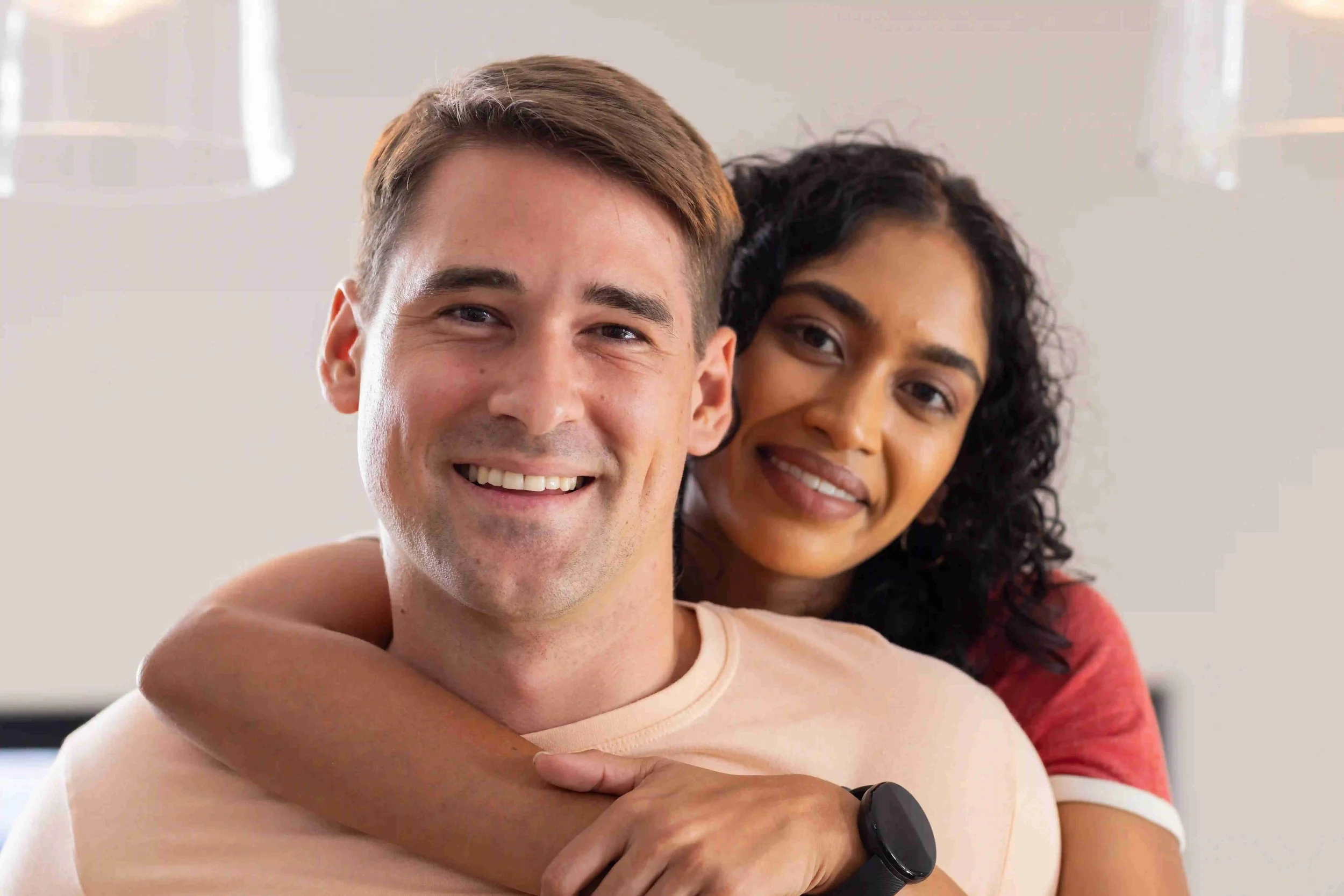 A smiling man with light skin and short brown hair being hugged from behind by a woman with darker skin and curly black hair, in a bright indoor setting.