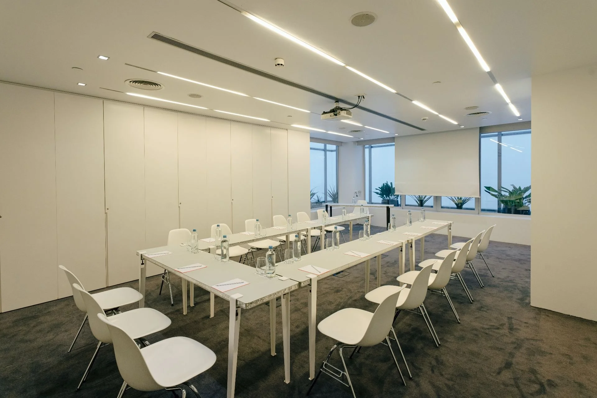 Empty conference room with white tables arranged in a U-shape, white chairs, water bottles, glassware, notepads, and pens. Large windows with a city view and potted plants. Overhead lighting and a projector mounted on the ceiling.