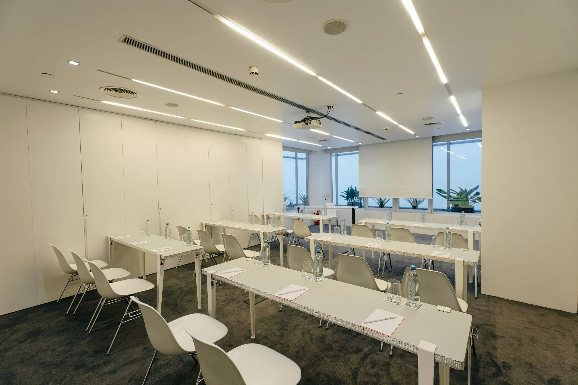 Empty conference room with white tables and chairs, water bottles, notepads, and glasses, illuminated by ceiling lights, with large windows and potted plants outside.
