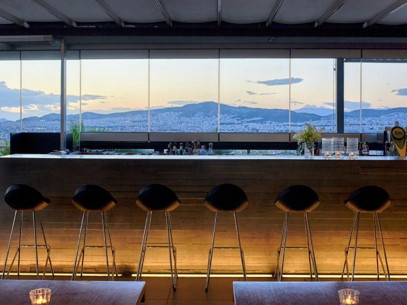 Modern bar with black barstools, a wooden bar counter, and a large window showing a cityscape and mountains at sunset.