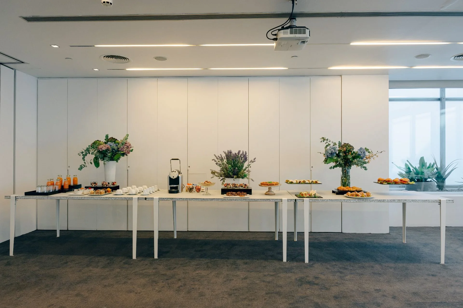 A buffet table with various pastries, finger foods, and beverages, decorated with three large floral arrangements in white vases, in a modern room with white walls and a projector mounted on the ceiling.