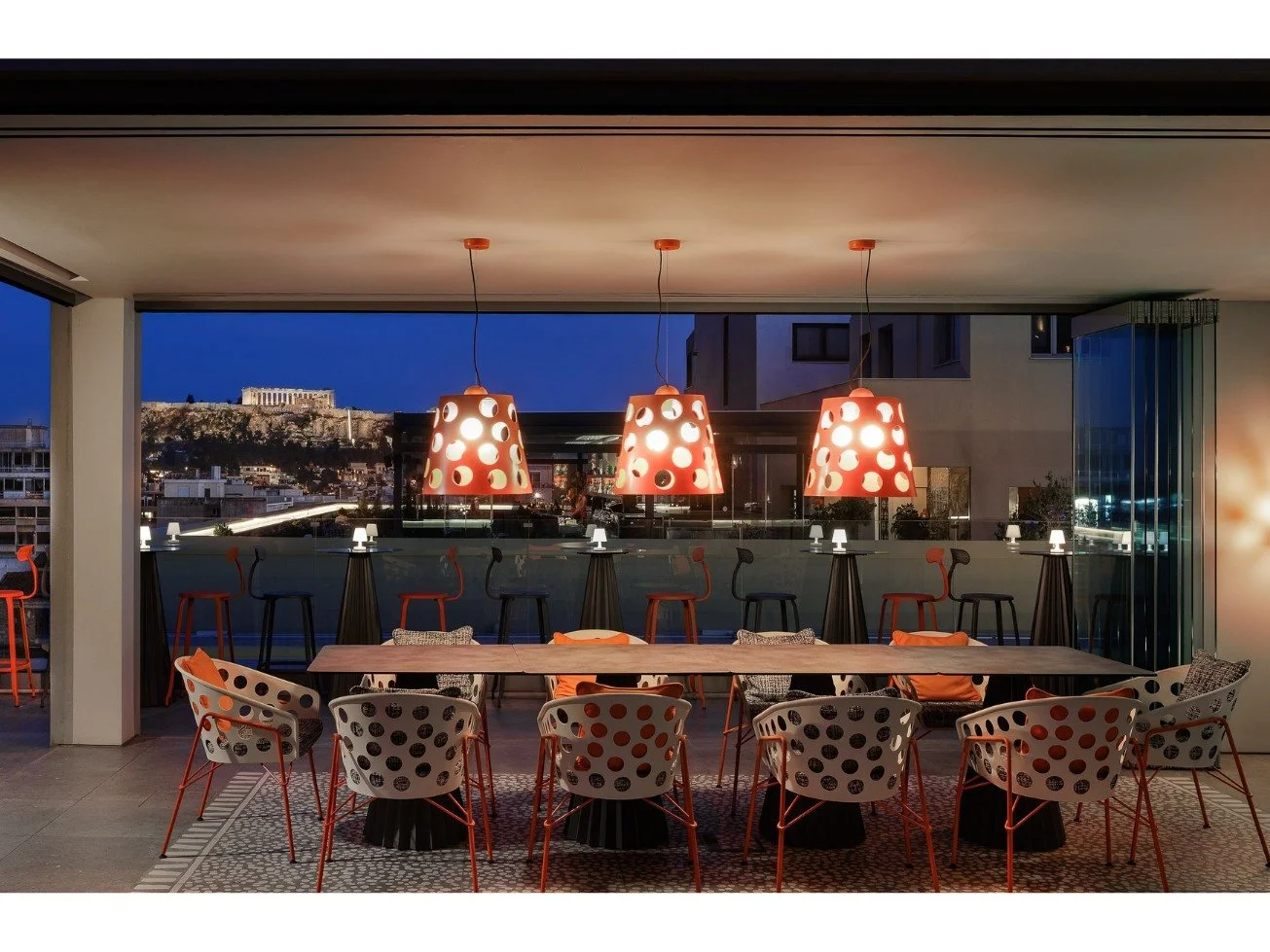 Modern dining area with a long wooden table, surrounded by white chairs with black and orange polka dot designs, illuminated by three orange pendant lights with circular cutouts, overlooking a cityscape at dusk.