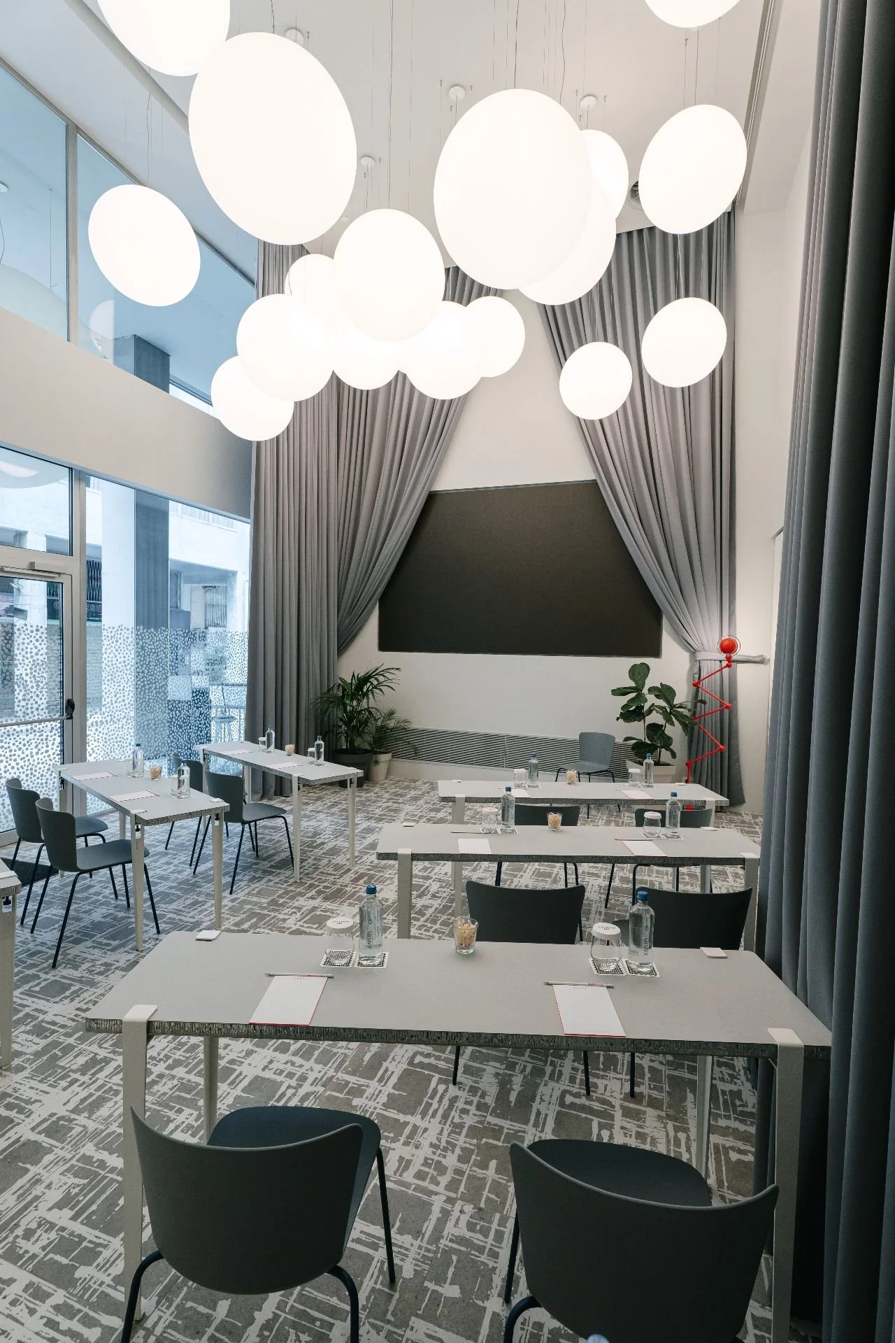 Modern conference room with tables arranged for a meeting, water bottles, notepads, and pens on each table, large gray curtains, tall windows, decorative lighting, potted plants, and a large blackboard or presentation screen on the wall.