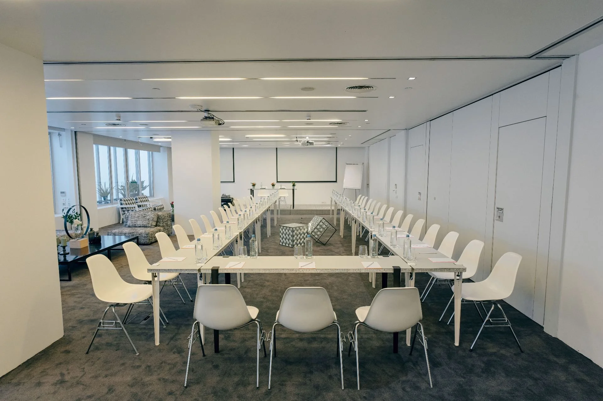Empty conference room with white tables arranged in a U-shape, white chairs, water bottles, notepads, pens, and glasses on tables, large windows with plants outside, decorated with plants and a black coffee table with a flower arrangement, a whiteboard, and a screen at the front.