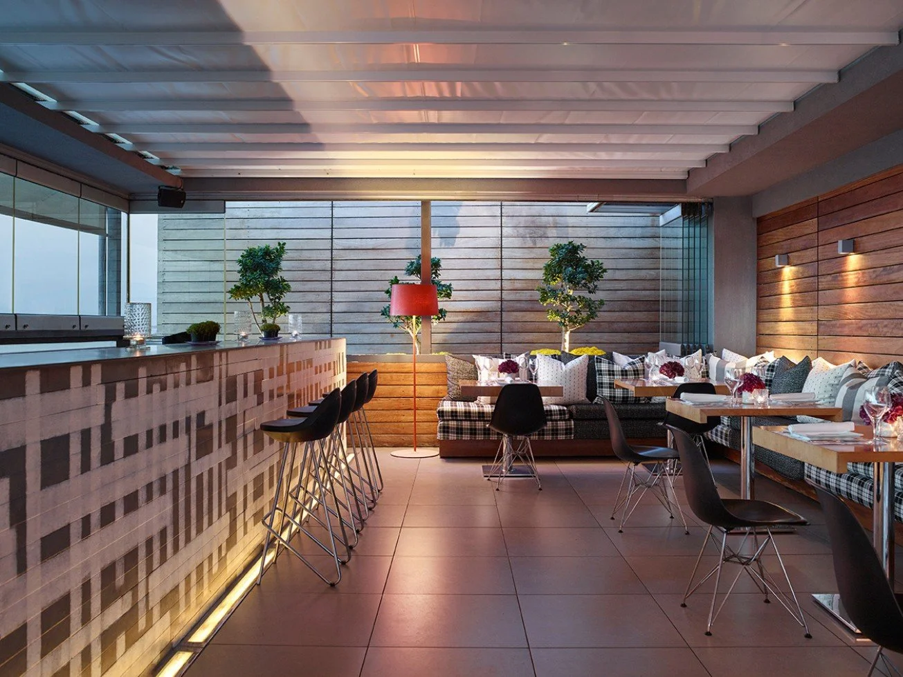 Modern restaurant interior with a bar counter, barstools, potted plants, a red floor lamp, and seating area with checkered and patterned cushions.