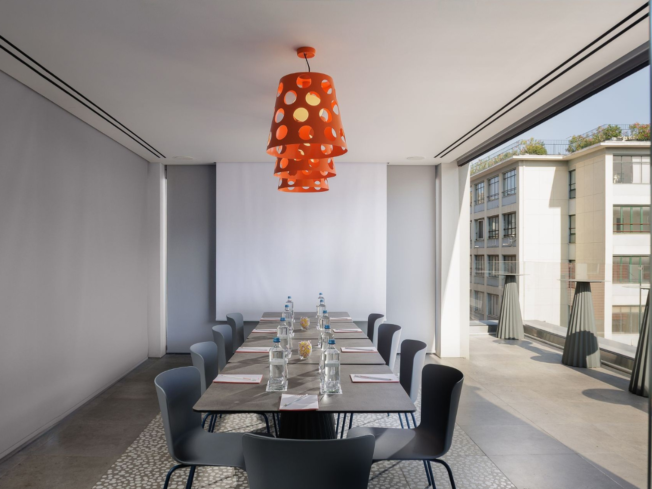 Modern conference room with a rectangular table set with water bottles, glasses, notepads, and flowers. Orange pendant lights hang from the ceiling. Large windows on one side offer a view of neighboring buildings.