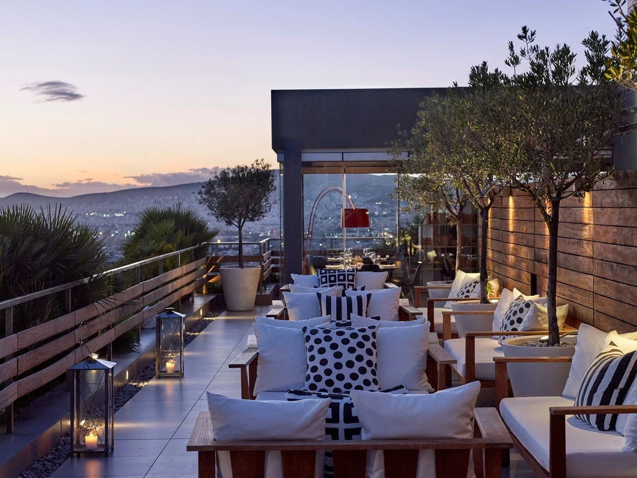 Outdoor rooftop patio with wooden furniture, pillows, trees, and lanterns, overlooking a cityscape at sunset.