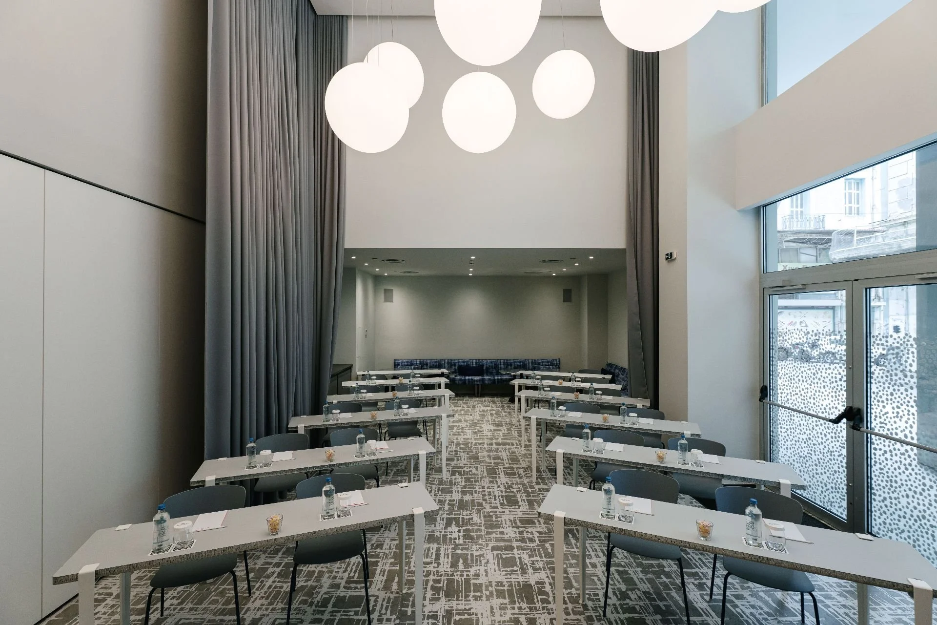 Conference room with white tables, black chairs, water bottles, and snacks, illuminated by large circular ceiling lights and natural light from tall windows.