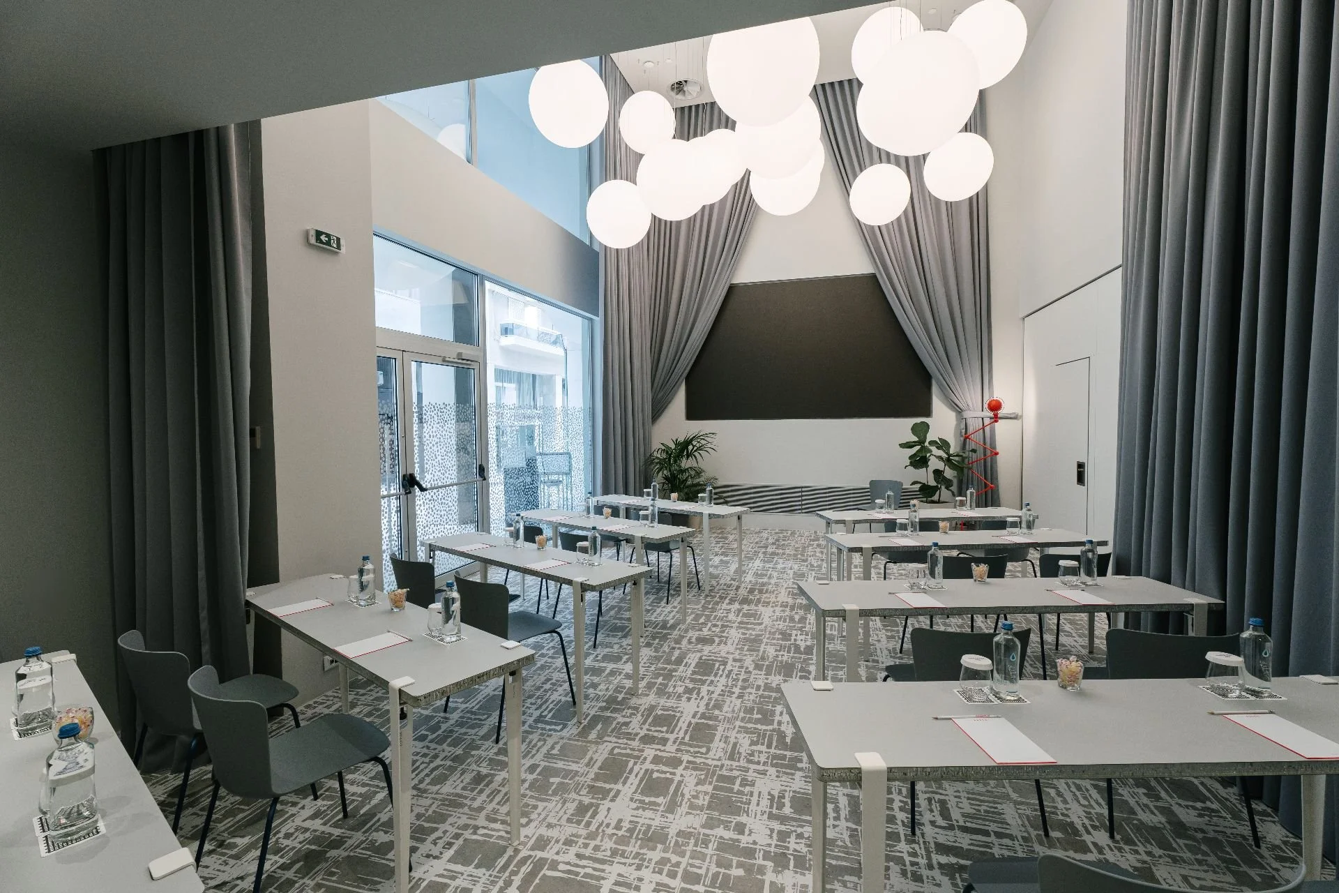 A conference room with rectangular tables set with water bottles, glasses, notepads, and pens, arranged in a U-shape facing a stage with a black screen, large gray curtains, potted plants, and modern pendant lights hanging from the ceiling.