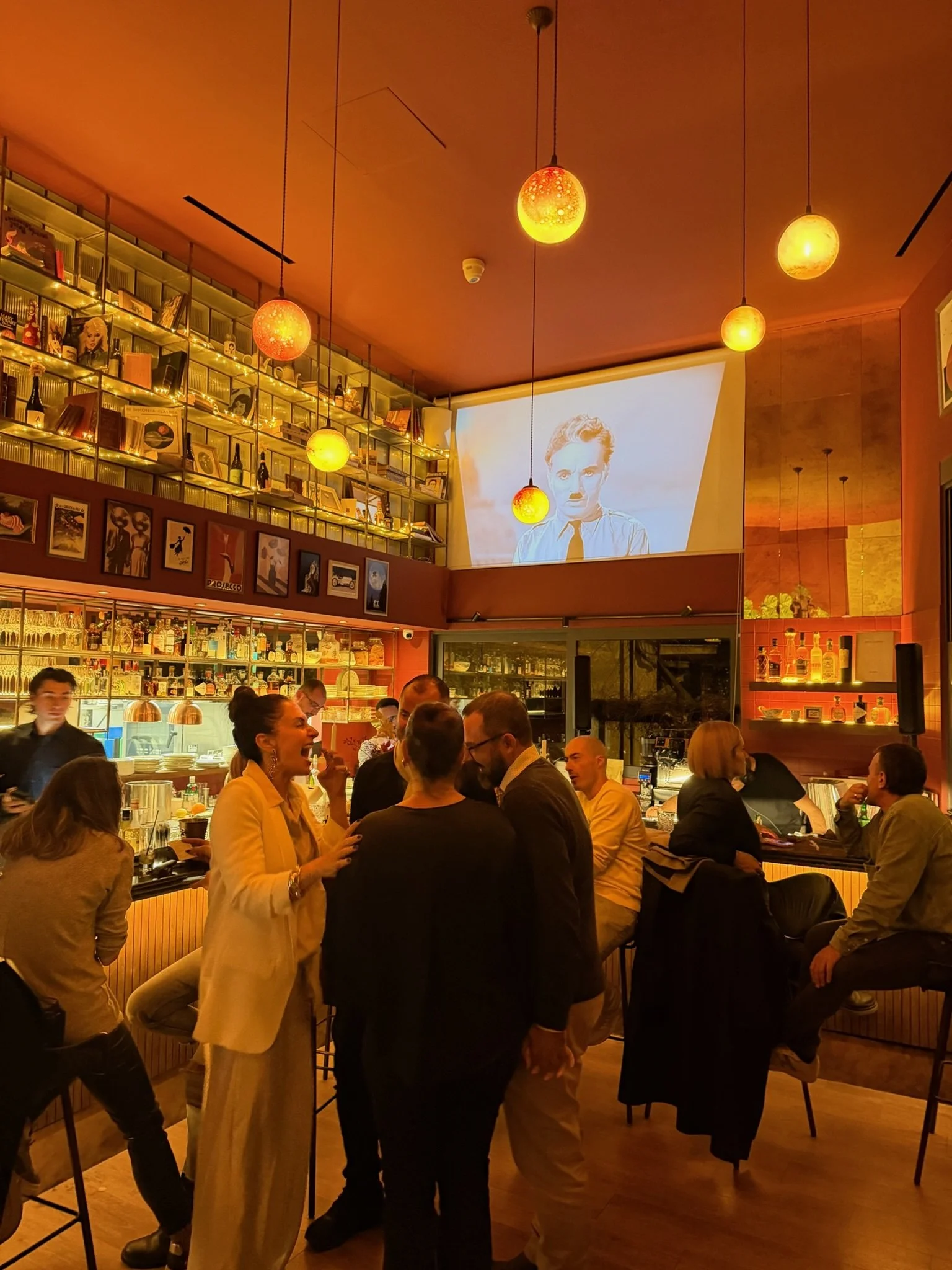 People gathered in a warmly lit bar or restaurant with shelves of bottles and decorations, watching a black-and-white video on a large screen.