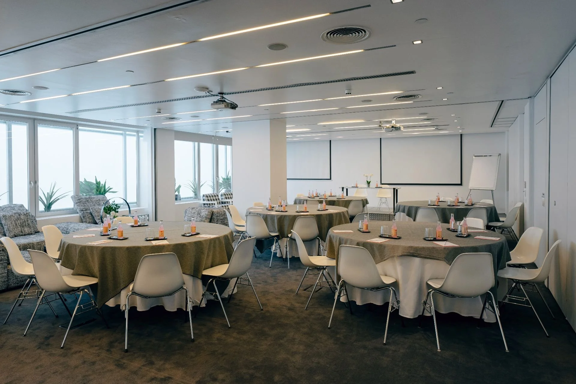 Conference room set up with round tables covered in beige tablecloths, each with bottled drinks and notepads, white chairs surrounding the tables, a whiteboard on a stand, and a projection screen on the wall, with large windows and indoor plants in the background.