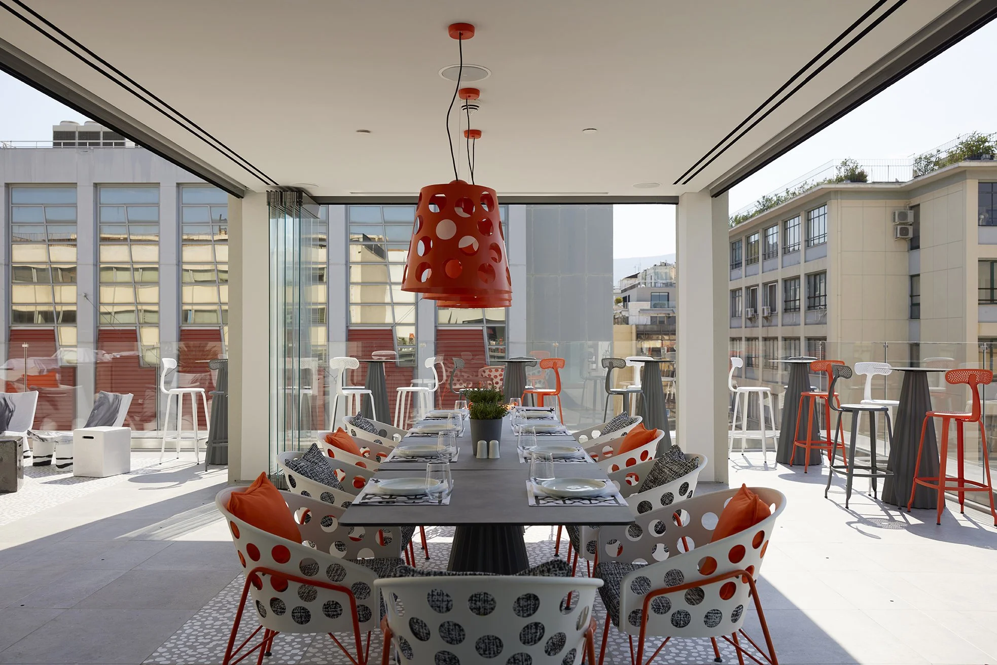 Interior of a modern restaurant with a long dining table set with plates, glasses, and napkins. Brightly colored chairs with orange and black cushions surround the table. Hanging orange pendant light fixtures above, with large windows letting in natural light, and an outdoor balcony with high stools in the background.