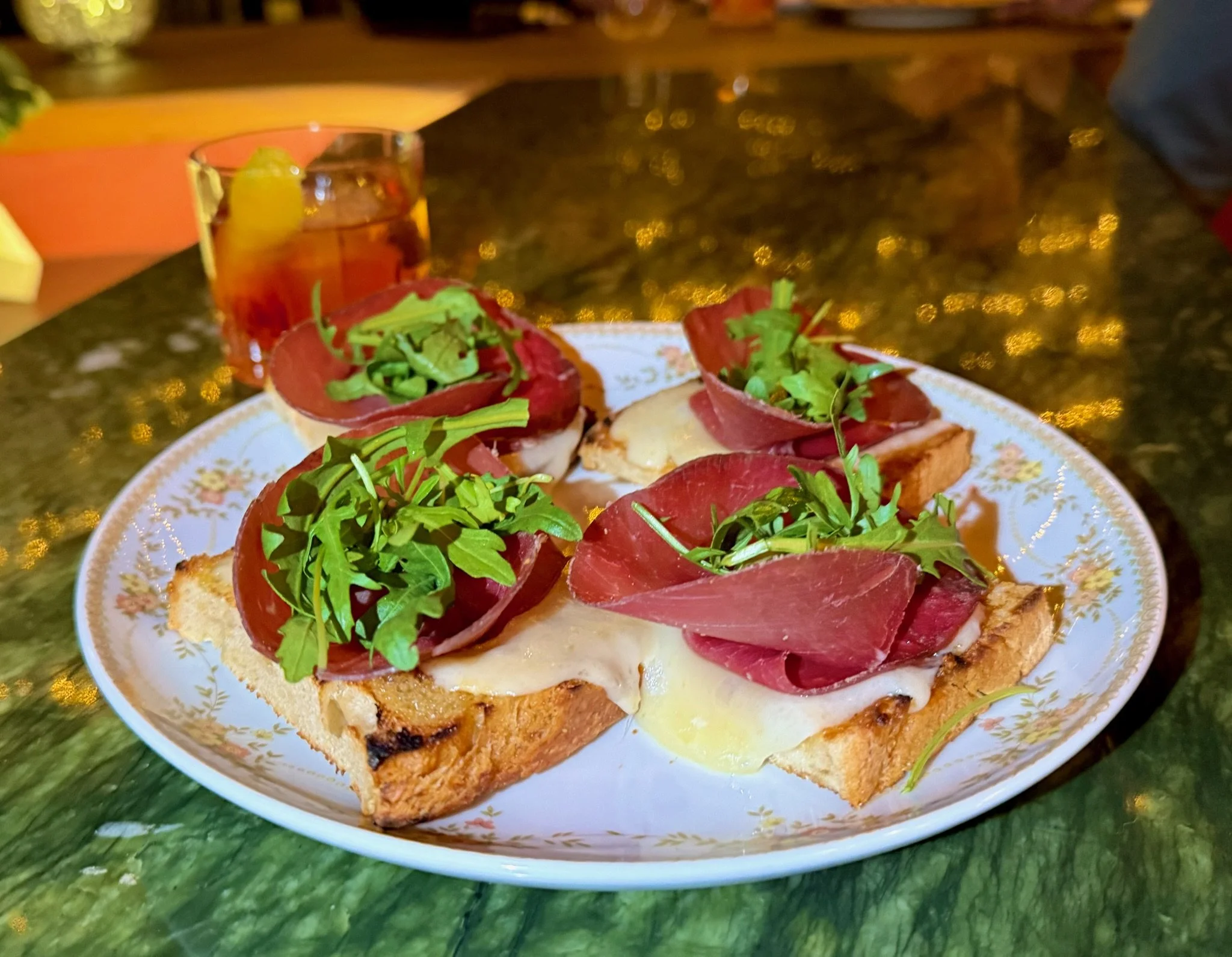 A plate with four slices of toasted bread topped with melted cheese, slices of cured meat, and garnished with fresh arugula. In the background, a glass of iced tea with lemon.