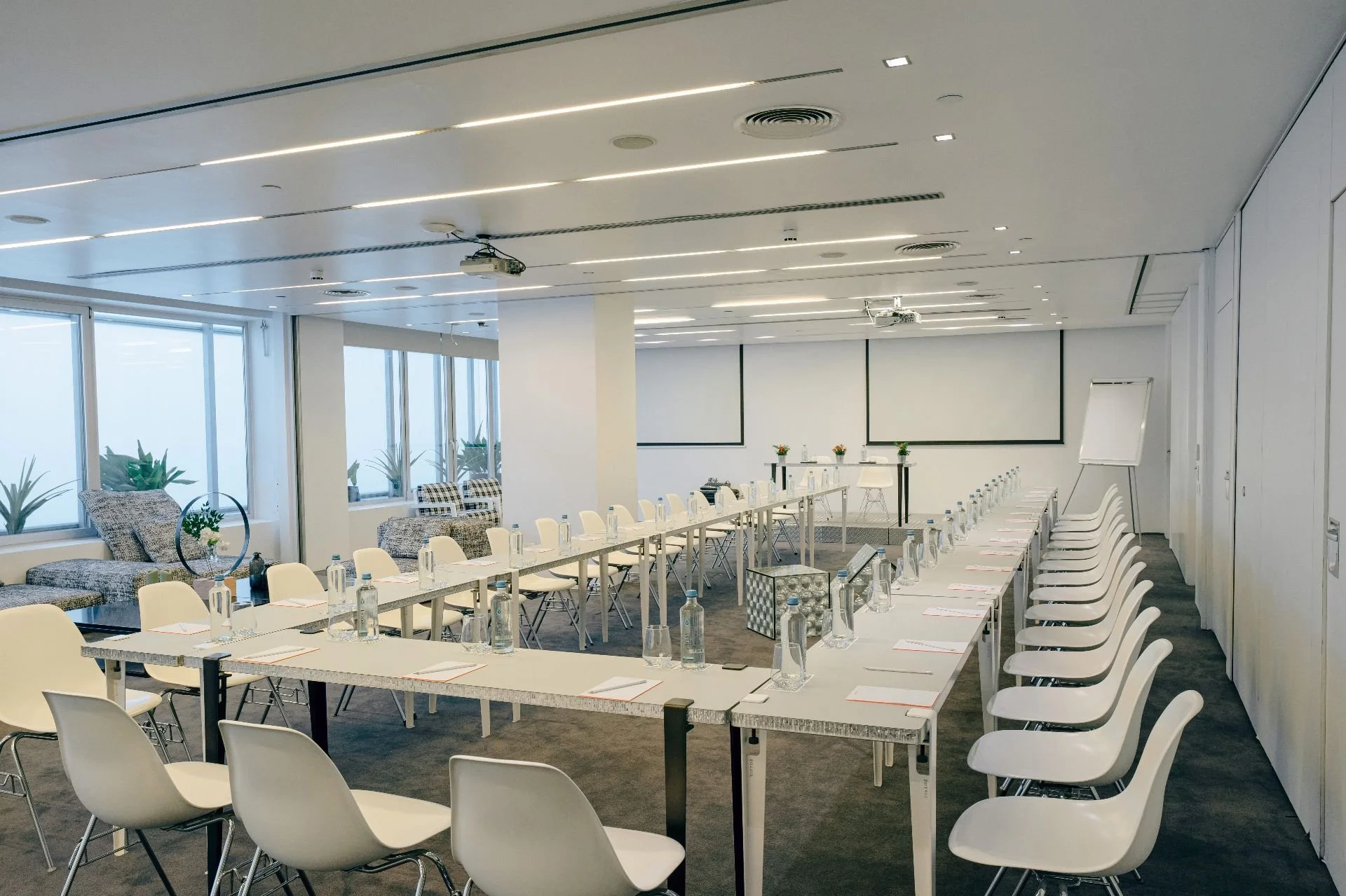 Empty conference room with U-shaped table setup, white chairs, large windows, and presentation screens at the front.