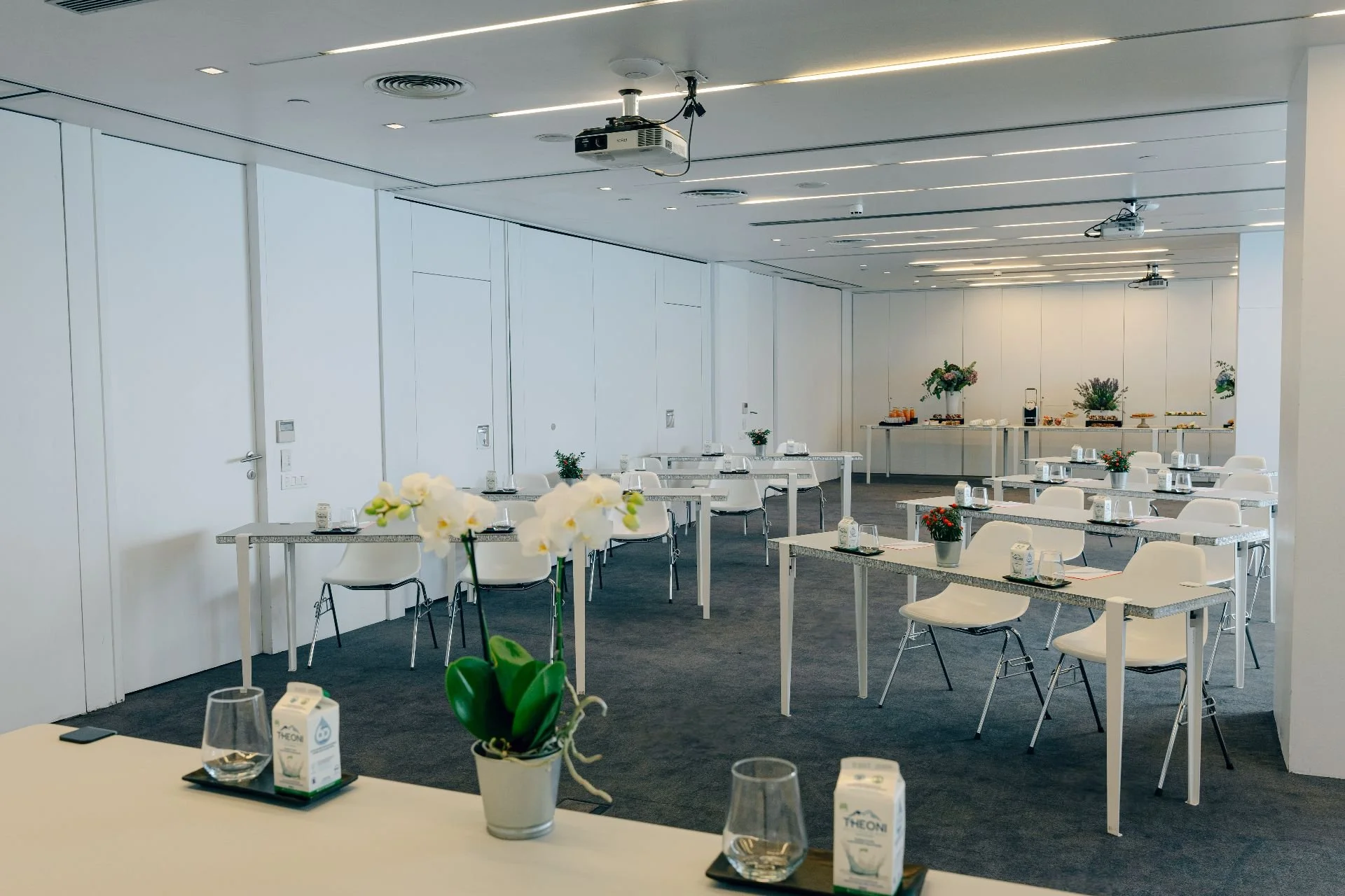 Empty conference room with white tables and chairs, decorated with potted flowers, with a refreshment table at the back, in a modern setting with minimal decor and ceiling projectors.