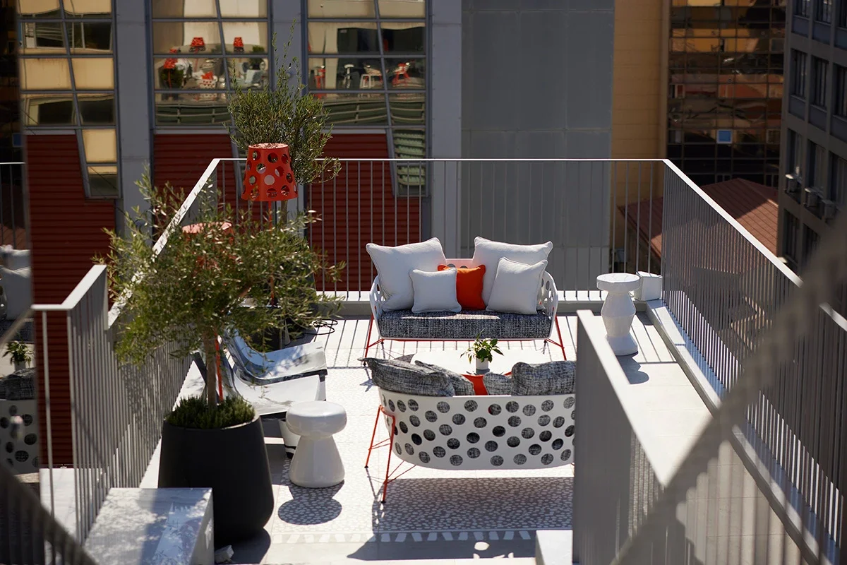 A rooftop balcony with modern furniture, including a circular lounge chair with polka dot pattern, a gray bench with cushions, white side tables, potted plants, and a decorative red and black lamp, with city buildings in the background.