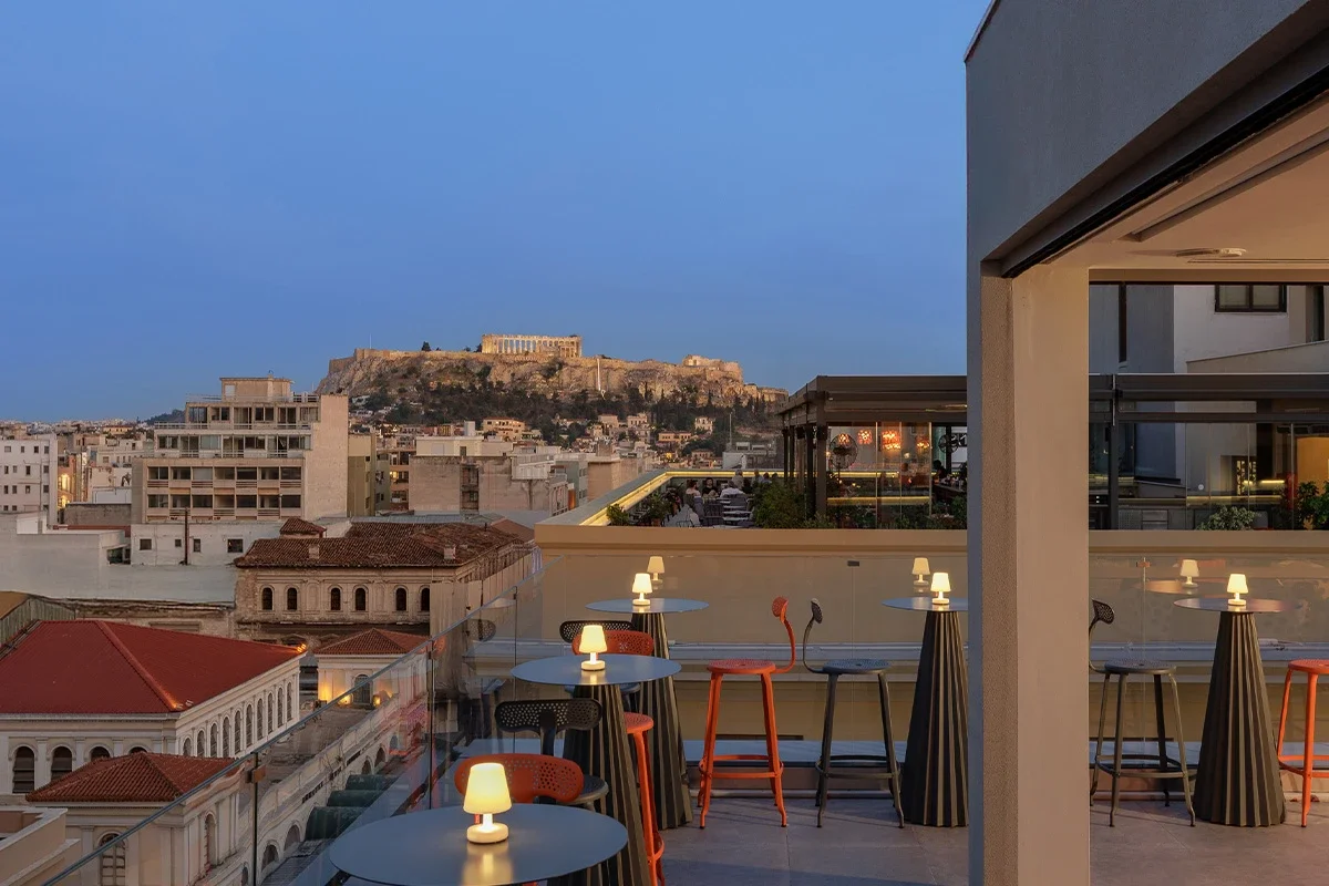 Rooftop bar with tables and chairs, small lamps on tables, overlooking the city with the Acropolis in the background, during dusk.