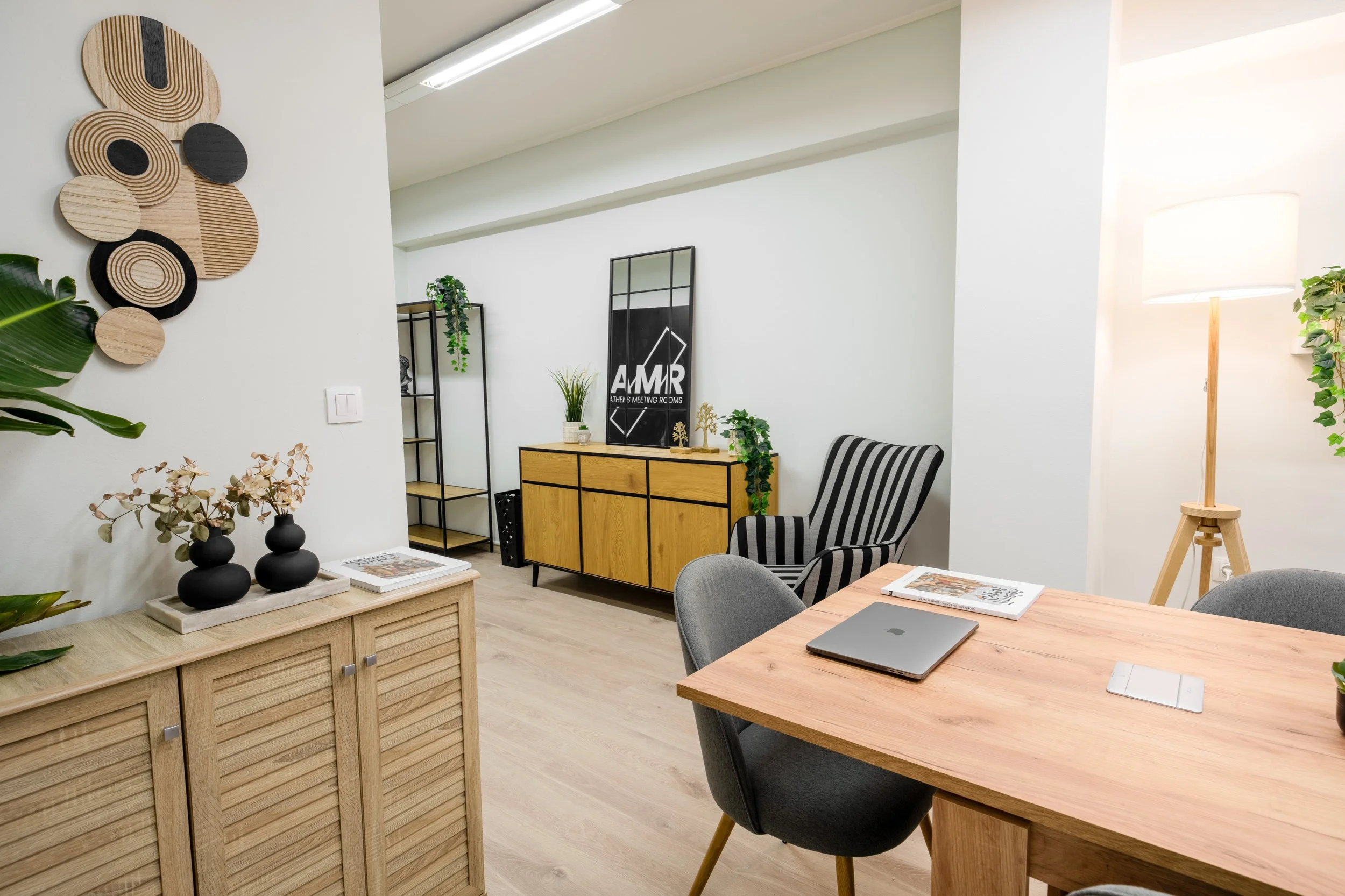 Modern living or office space with wooden furniture, decorative plants, and wall art, featuring a wooden dining table with a closed laptop, magazines, and a wireless mouse, illuminated by a standing lamp, with light-colored flooring and white walls.