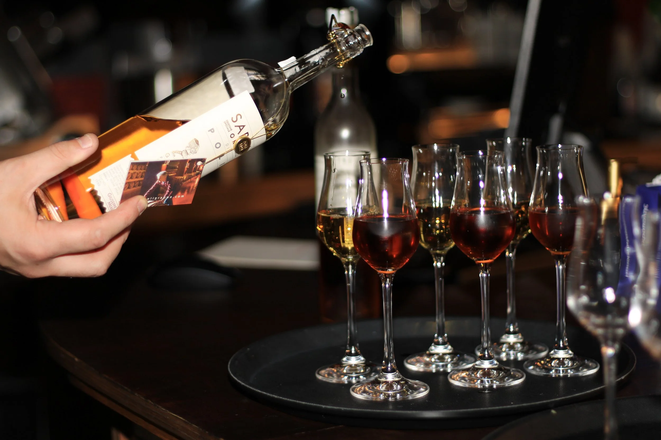 A person pouring a bottle of rosé wine into glasses on a black tray on a wooden table at a bar or restaurant.