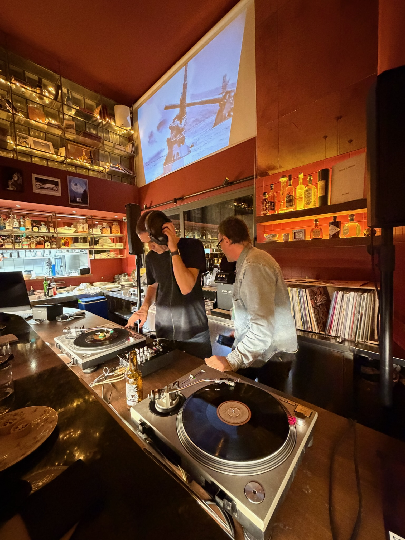 Two DJs working at a bar with turntables, music equipment, and a large screen displaying a helicopter scene.