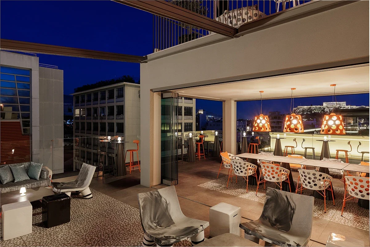 Nighttime view of a modern rooftop bar with seating areas, hanging orange pendant lights, and city buildings in the background.