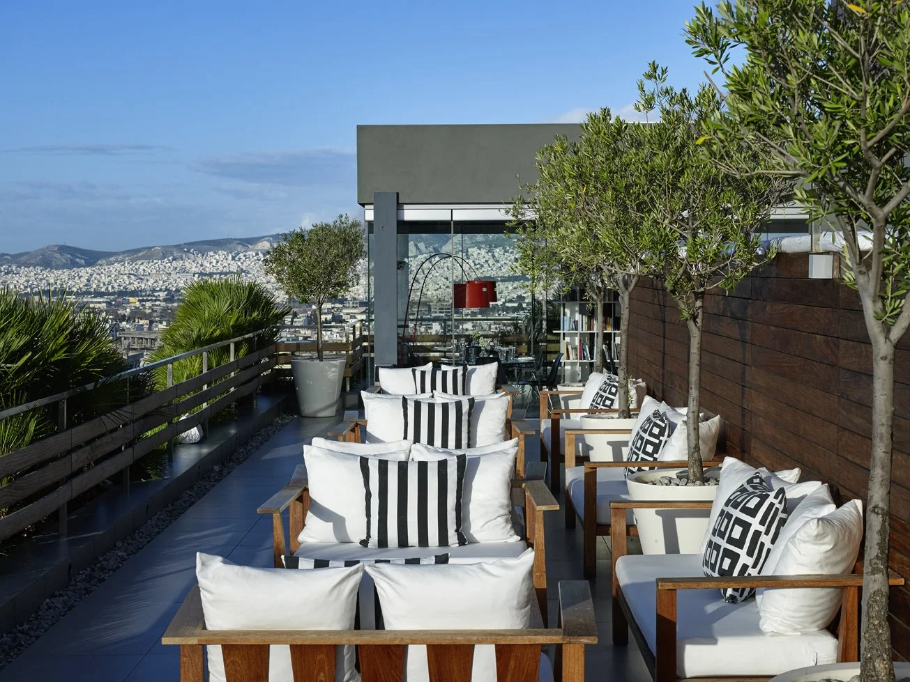 Rooftop patio with wooden chairs with white cushions and black-and-white patterned pillows, potted trees, and cityscape view in the background under a clear sky.