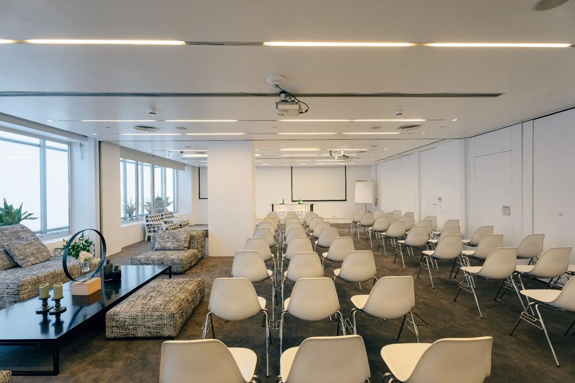 Empty conference room with rows of white chairs facing a projection screen, with a seating area on the left and windows along the left wall.
