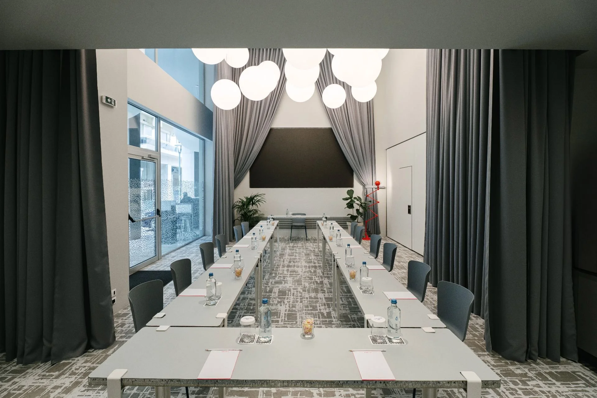 A modern conference room with U-shaped tables, chairs, and large windows with curtains, decorated with ceiling lights, plants, and a chalkboard.