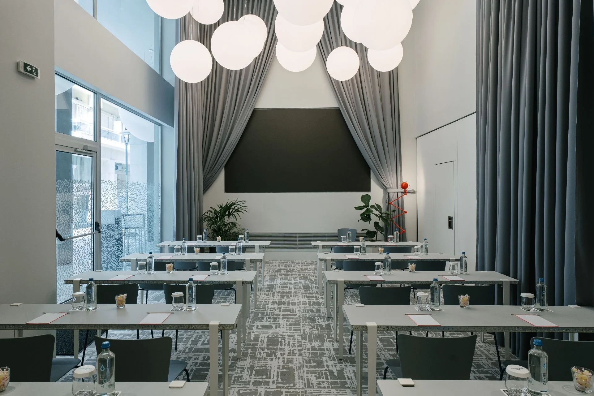 Conference room with white tables, black chairs, water bottles, notepads, and glasses arranged for a meeting, large gray curtains, a black chalkboard, potted plants, and modern light fixtures hanging from the ceiling.