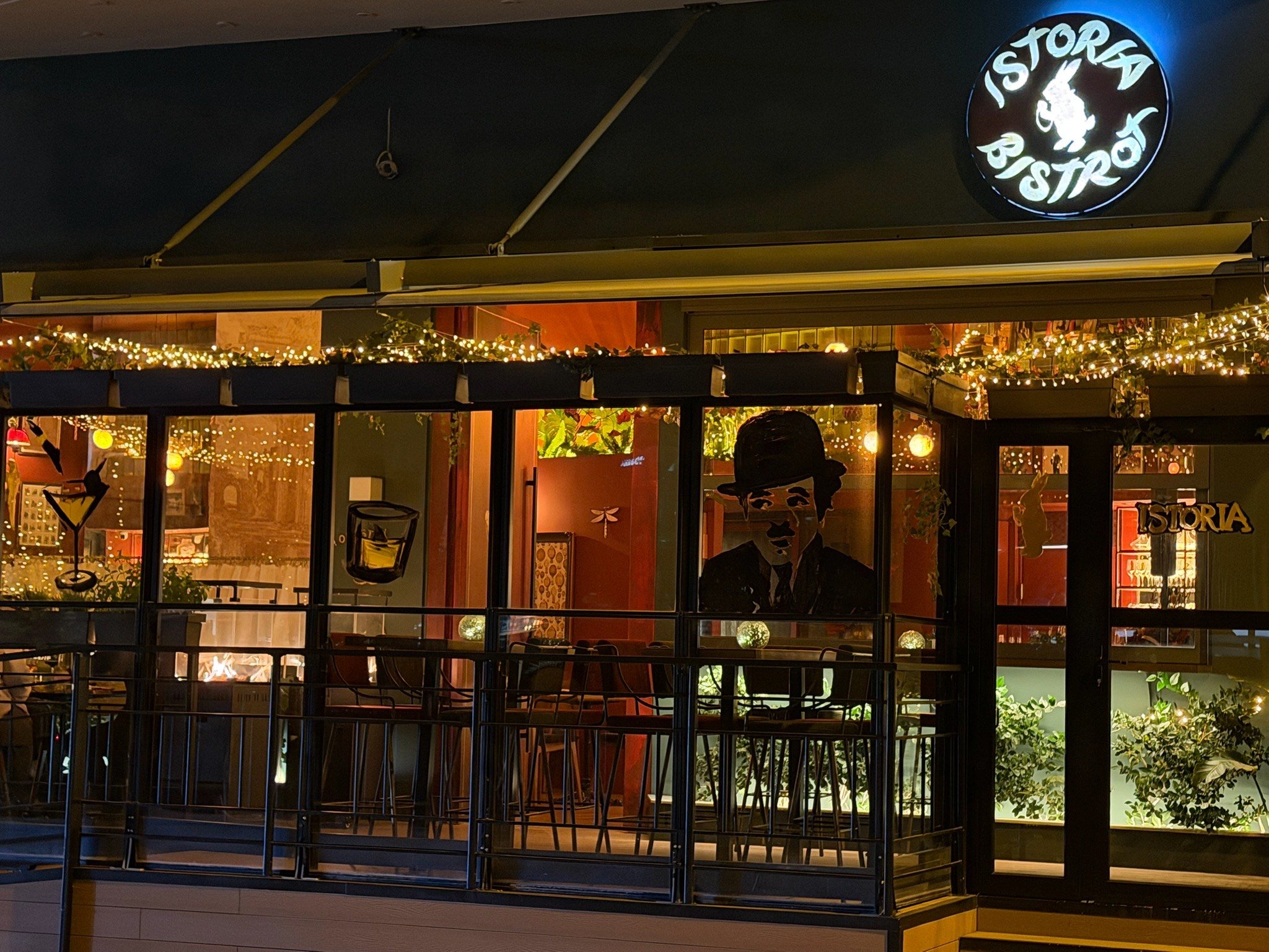 Night view of a cozy restaurant named 'Istoria Bistro' with warm yellow and orange lighting, decorated with string lights and greenery, and a silhouette of a waiter painted on the window.