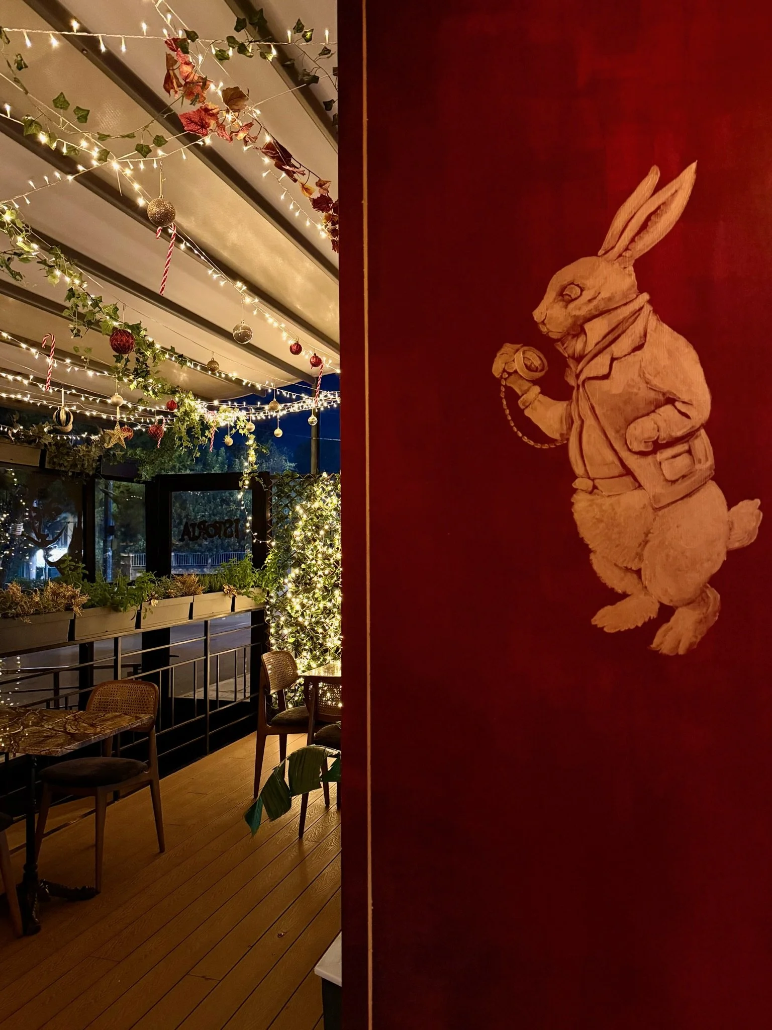 A cozy indoor space decorated with string lights, ornaments, and greenery hanging from the ceiling. There are a few chairs and a table on a wooden floor, and a large window in the background. A wooden sculpture of a rabbit dressed in clothing, holding a pocket watch, is on a red wall on the right side.