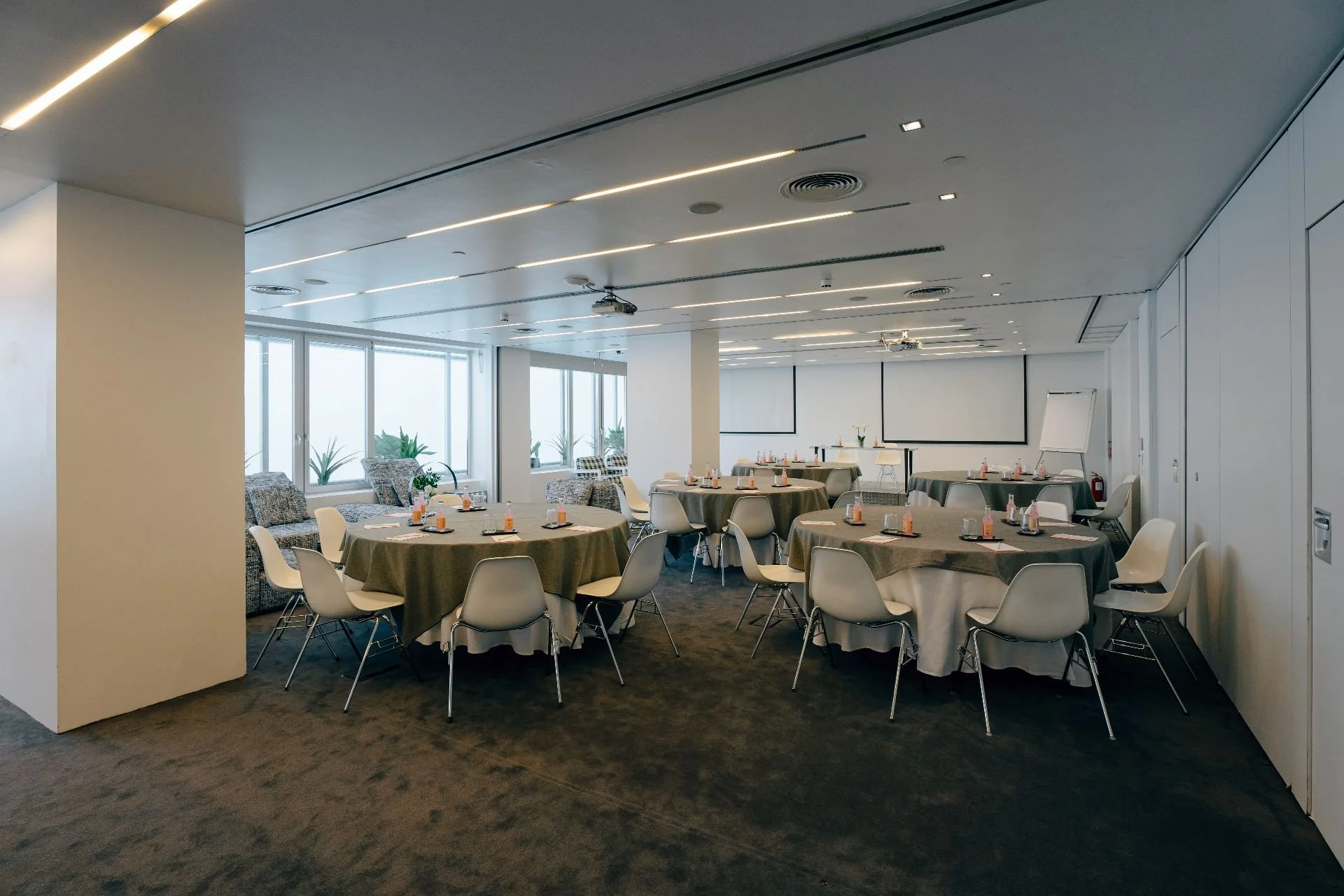 Conference room with round tables, chairs, a projector, whiteboards, and large windows with plants outside.
