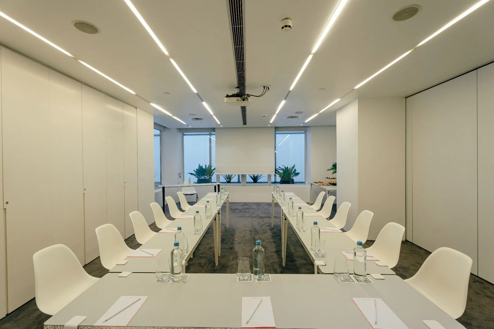 A modern, well-lit conference room with two rows of white chairs facing a central table, each with a notepad, pen, and bottled water. Windows with potted plants in the background, and a projector mounted on the ceiling.