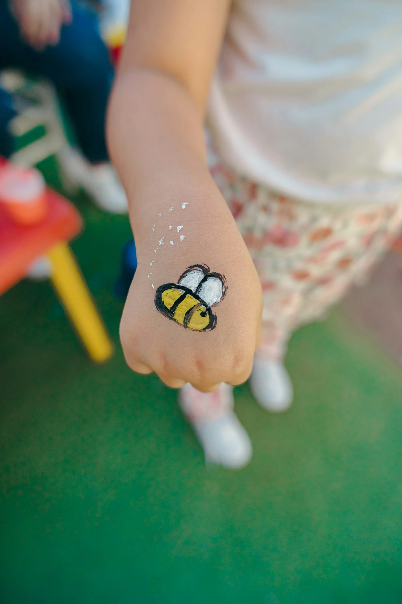 Child's hand with a painted bee on the back of the hand and small white dot details, with children and play area in the blurred background.