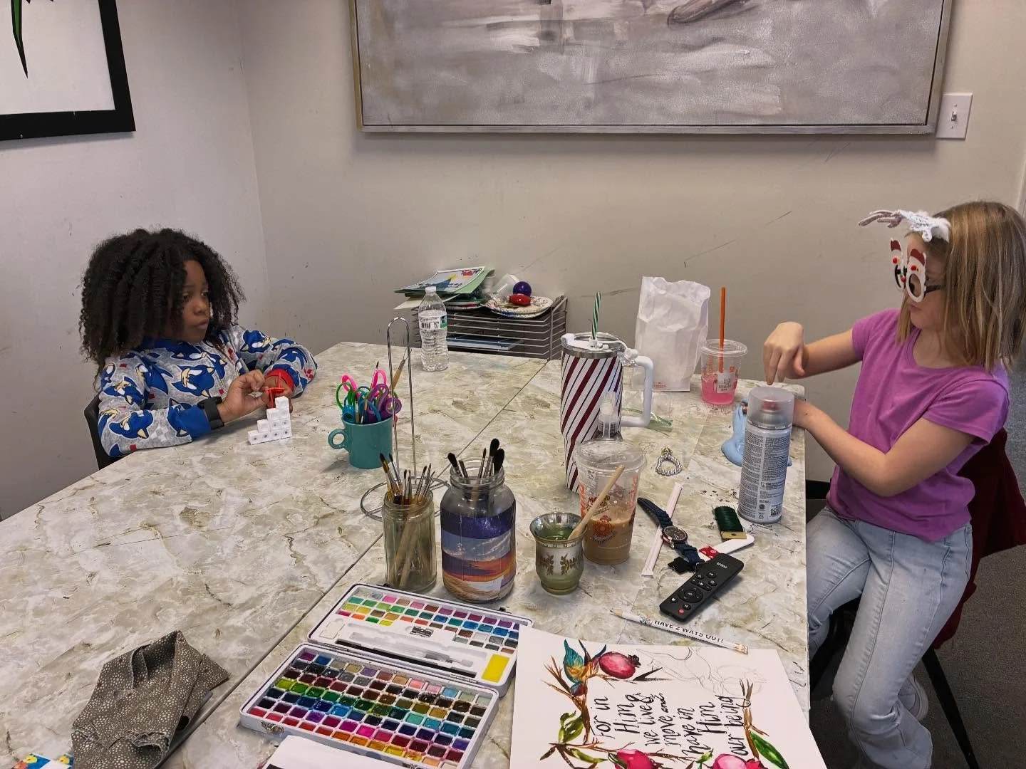 Two young girls sitting at a cluttered table engaging in arts and crafts. One girl has curly hair, wearing a colorful patterned shirt, holding a small craft object. The other girl has straight blonde hair, glasses, a headband with decorations, and a pink shirt, is reaching for supplies. The table is scattered with cups, water bottles, art supplies, and a watercolor paint set. There is a large holiday-themed sign or poster with colorful flowers and a quote partially visible.