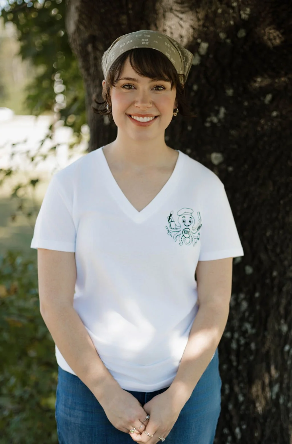 A young woman with short brown hair, wearing a beige bandana, white V-neck T-shirt with a logo, and blue jeans, standing outdoors near a tree, smiling at the camera.