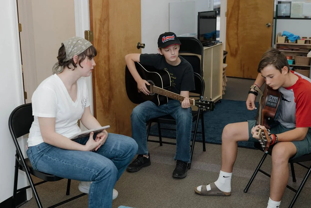 Three young people are sitting in a room, two of them playing guitars and one holding a tablet, possibly in a music lesson or casual jam session.