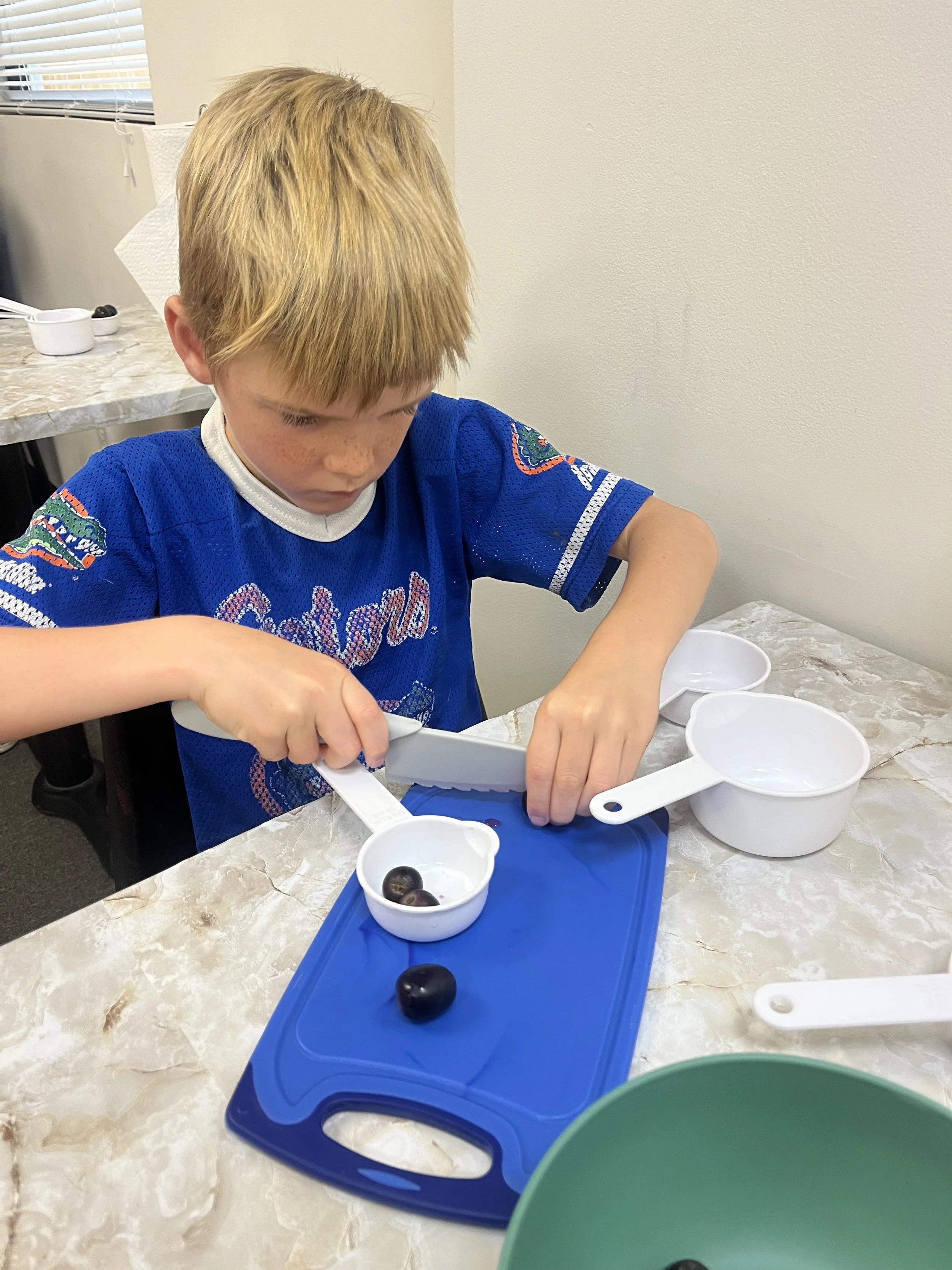 A young boy with blonde hair and freckles, wearing a blue sports jersey, is cutting blueberries on a blue cutting board with a plastic knife in a kitchen.