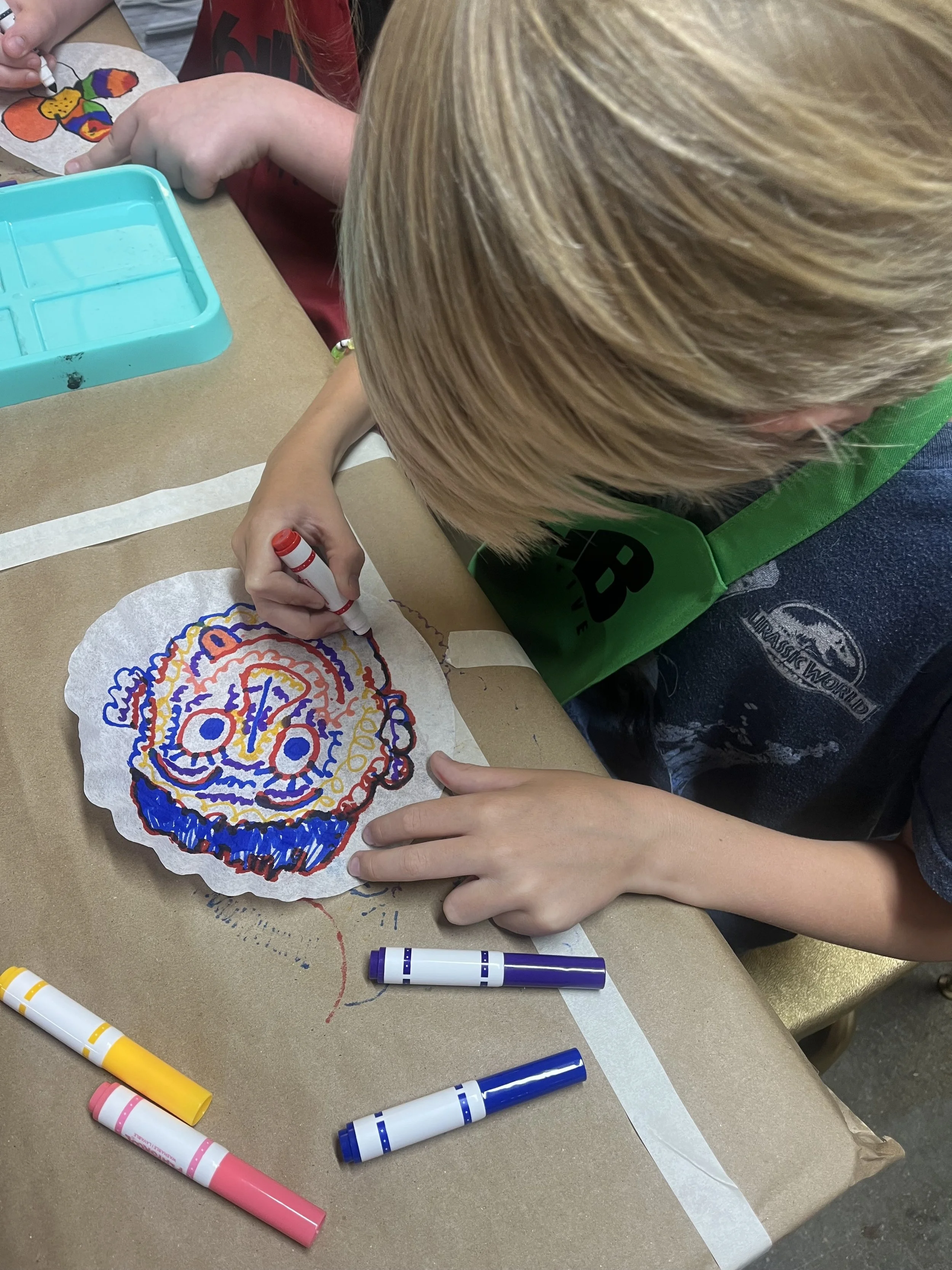 Child drawing a colorful face on a paper plate using markers, with art supplies on the table.