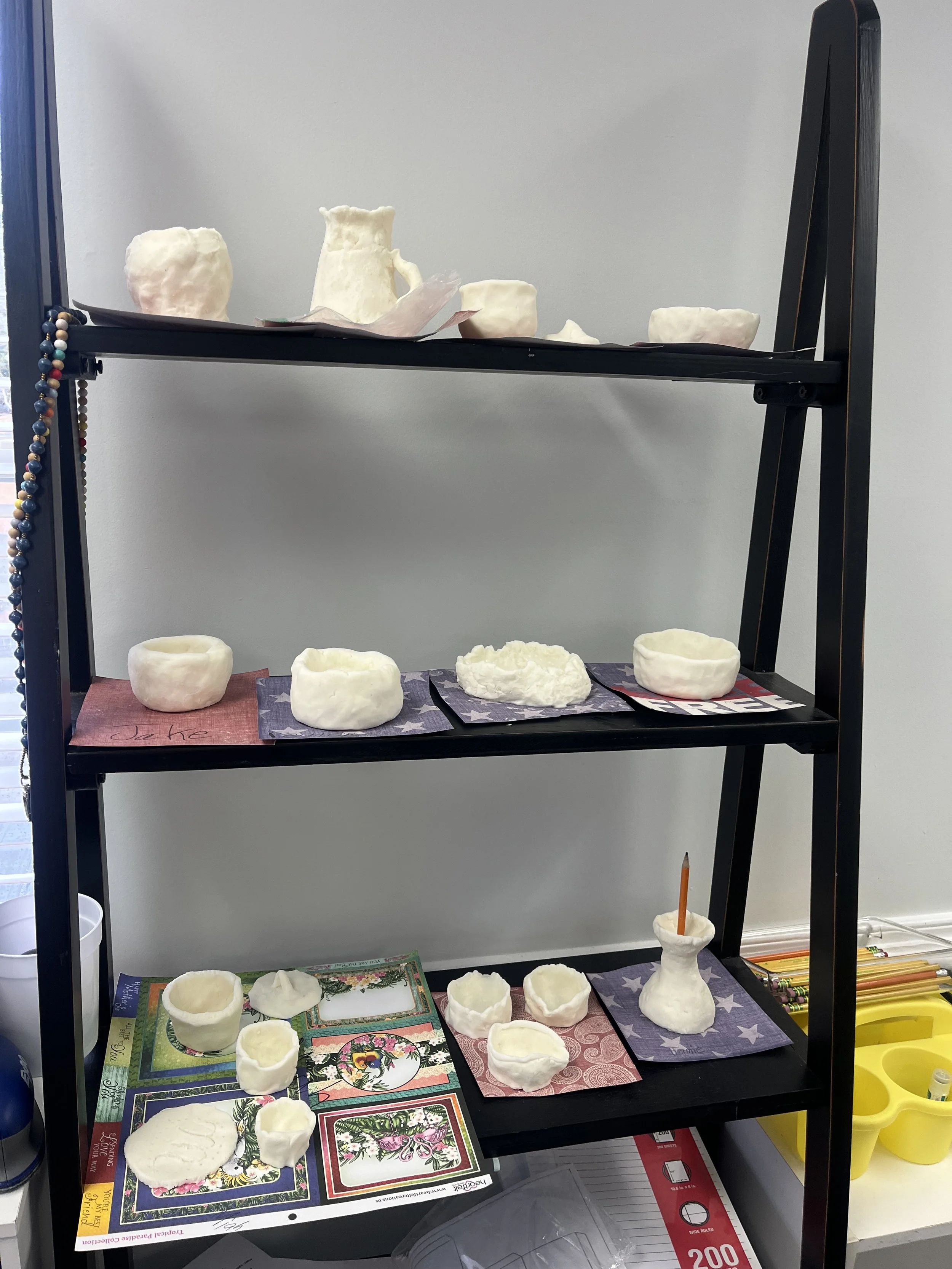Shelf displaying various white ceramic or plaster bowls and vessels at different stages of creation, with some placed on decorative paper or napkins.