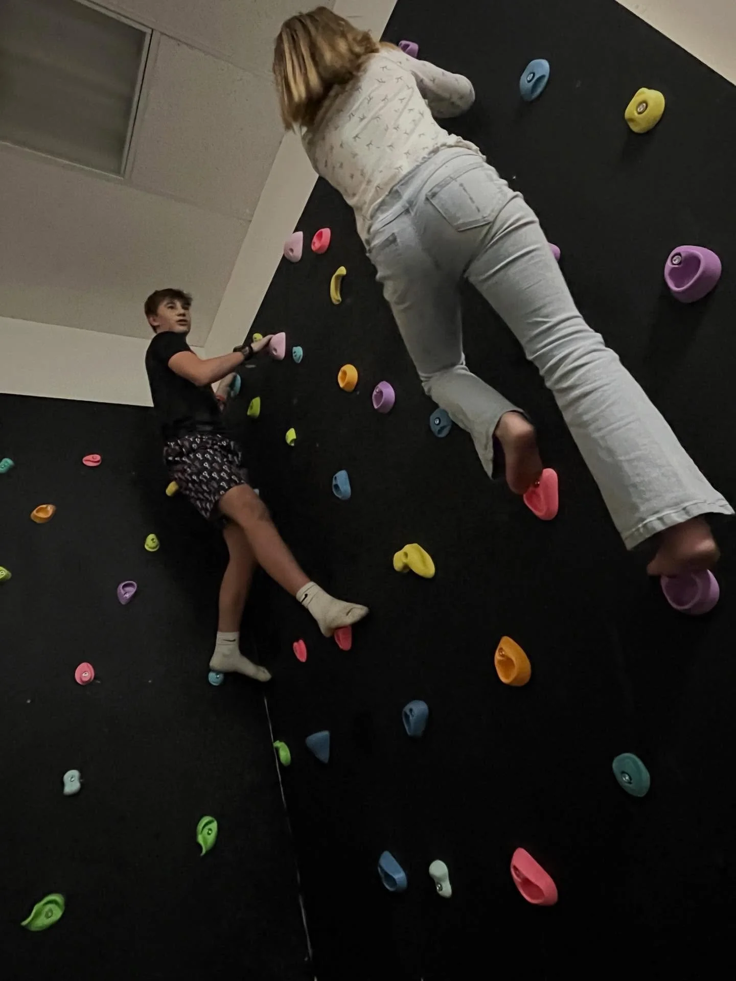 Two children climbing a colorful indoor rock wall with various brightly colored holds.