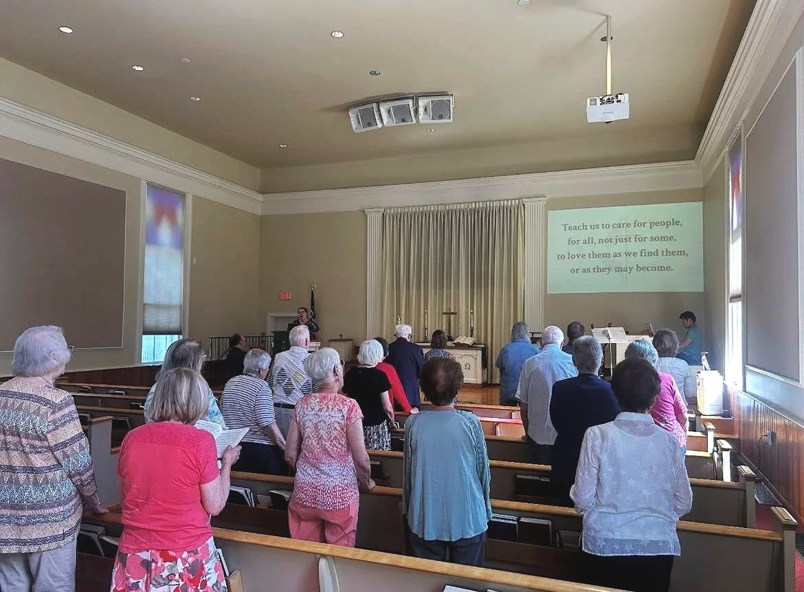 A person making a snow cone with a red flavored syrup, using a metal scoop, at an outdoor stall with jars of syrup in the foreground.