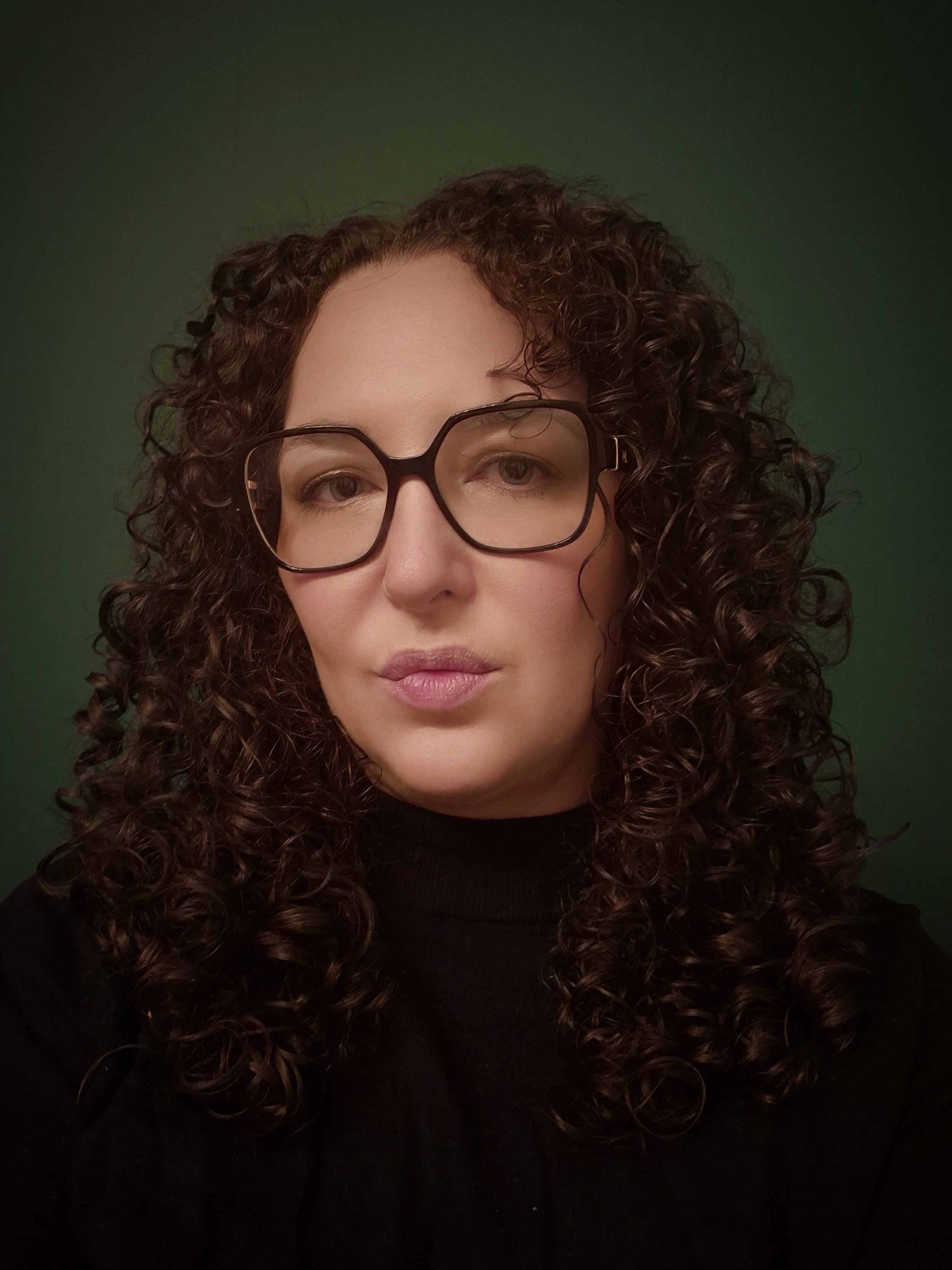 Studio-style portrait of a woman with long dark curly hair and glasses, wearing a black top, looking slightly toward the camera against a dark green background.