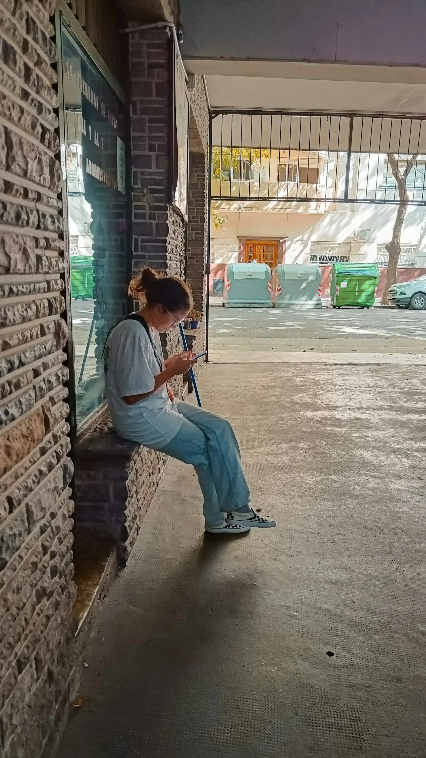 Young woman sitting outside against a wall, looking down at her phone.